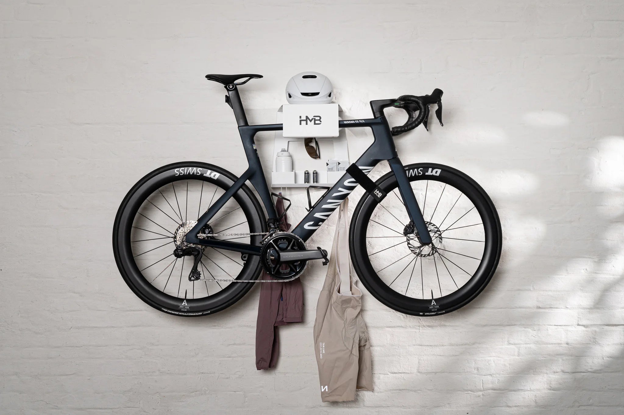 A wide lifestyle shot of a black Canyon road bike organized on a white HMB Ultimate wall mount, featuring a helmet on the top shelf, modular storage bins for nutrition and bottles, and a specialized HMB wheel strap securing the front wheel.