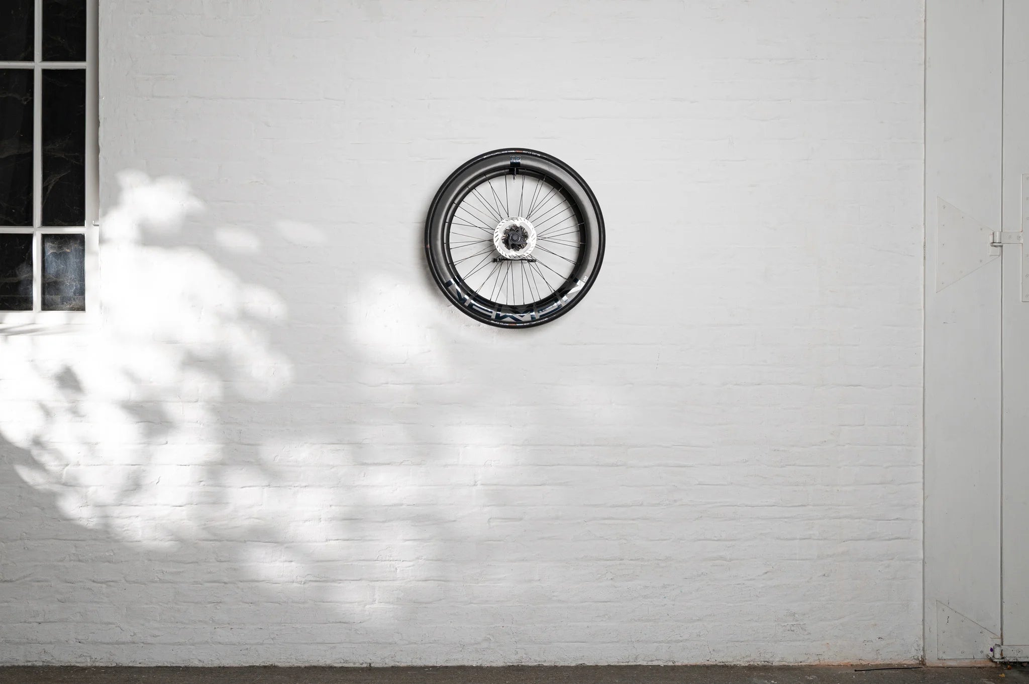 A high-quality carbon bike wheel securely and horizontally displayed on a white brick wall using the minimalist Hold My Bike (HMB) wheel mount for indoor storage.