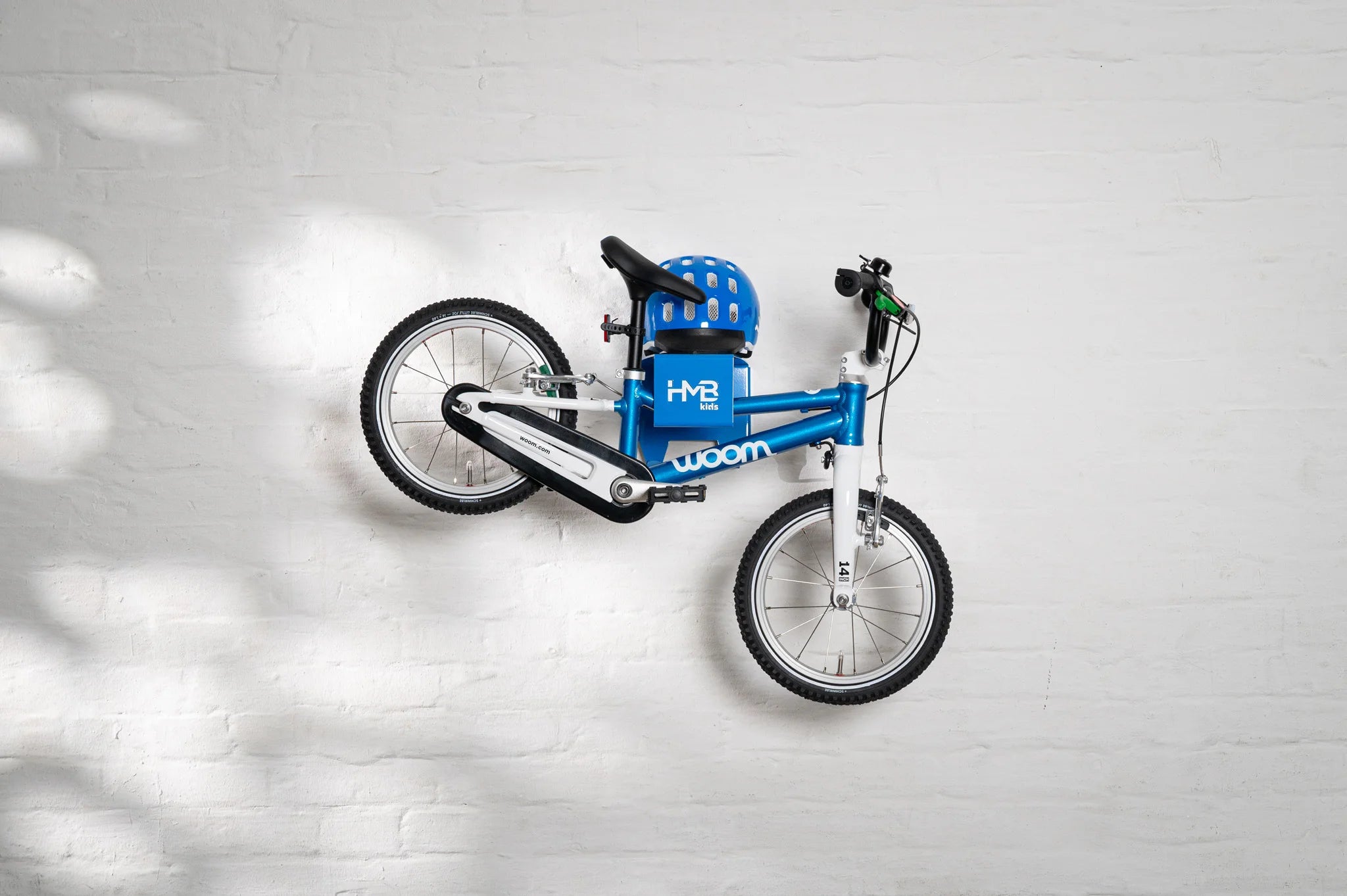 A blue Hold My Bike (HMB) Kids wall mount securely holding a blue woom 2 children's bike by the frame, with a matching blue woom helmet stored on the integrated top shelf.