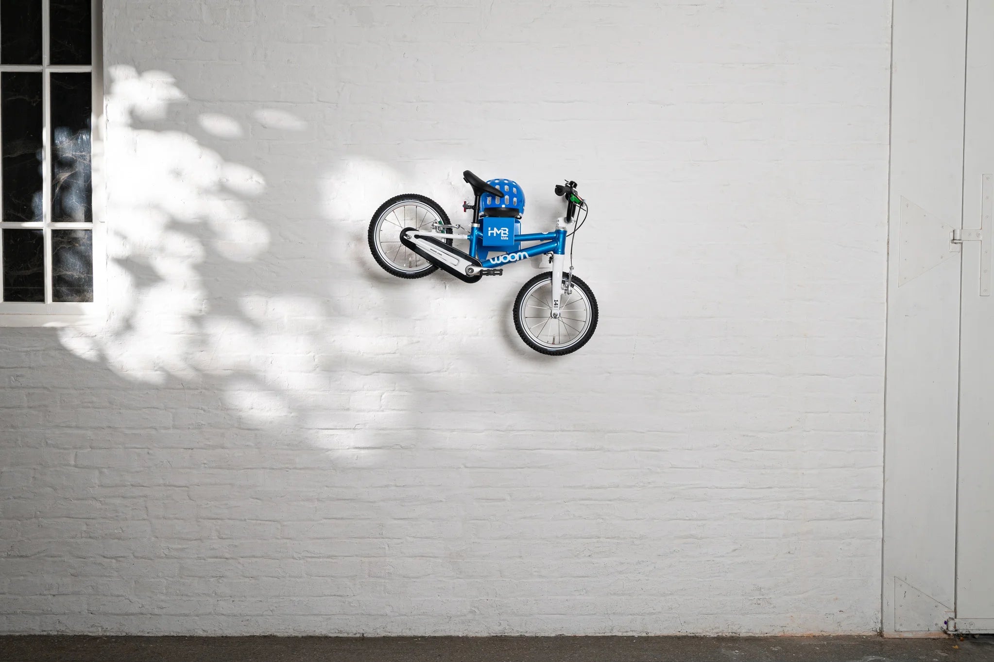 A blue Hold My Bike (HMB) Kids wall mount securely holding a blue woom 2 children's bicycle by the frame, with a matching blue woom helmet stored on the integrated top shelf against a white brick wall.