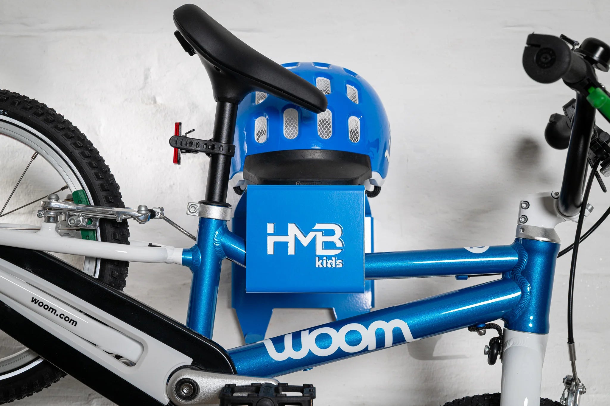 Close-up of a blue Hold My Bike (HMB) Kids wall mount holding a blue woom 2 bicycle by the frame, with the saddle positioned just above a matching blue woom helmet resting on the integrated top shelf.