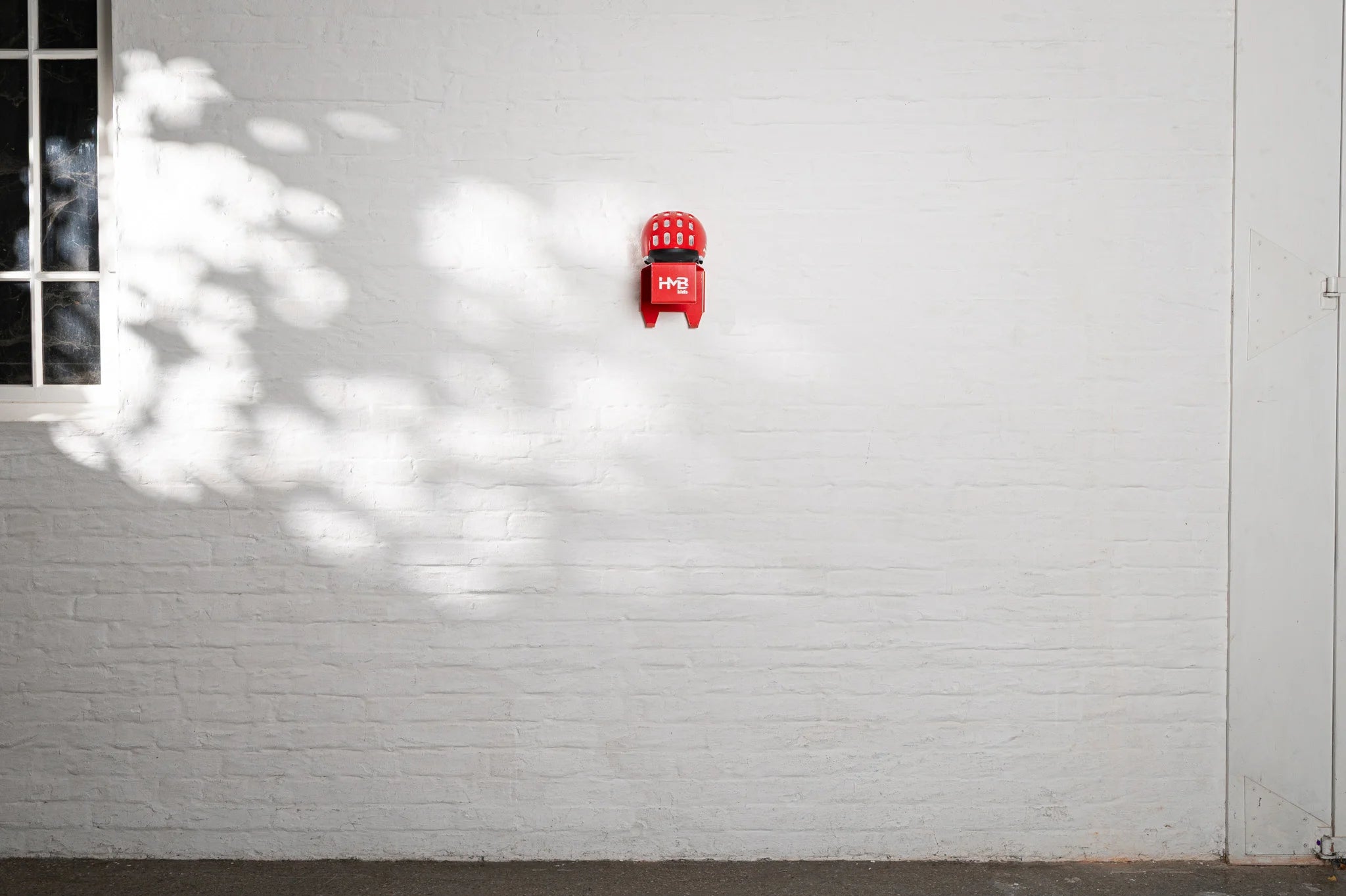A wide shot of a minimalist white brick wall featuring a single red Hold My Bike (HMB) Kids wall mount with a matching red woom helmet stored neatly on the top shelf.