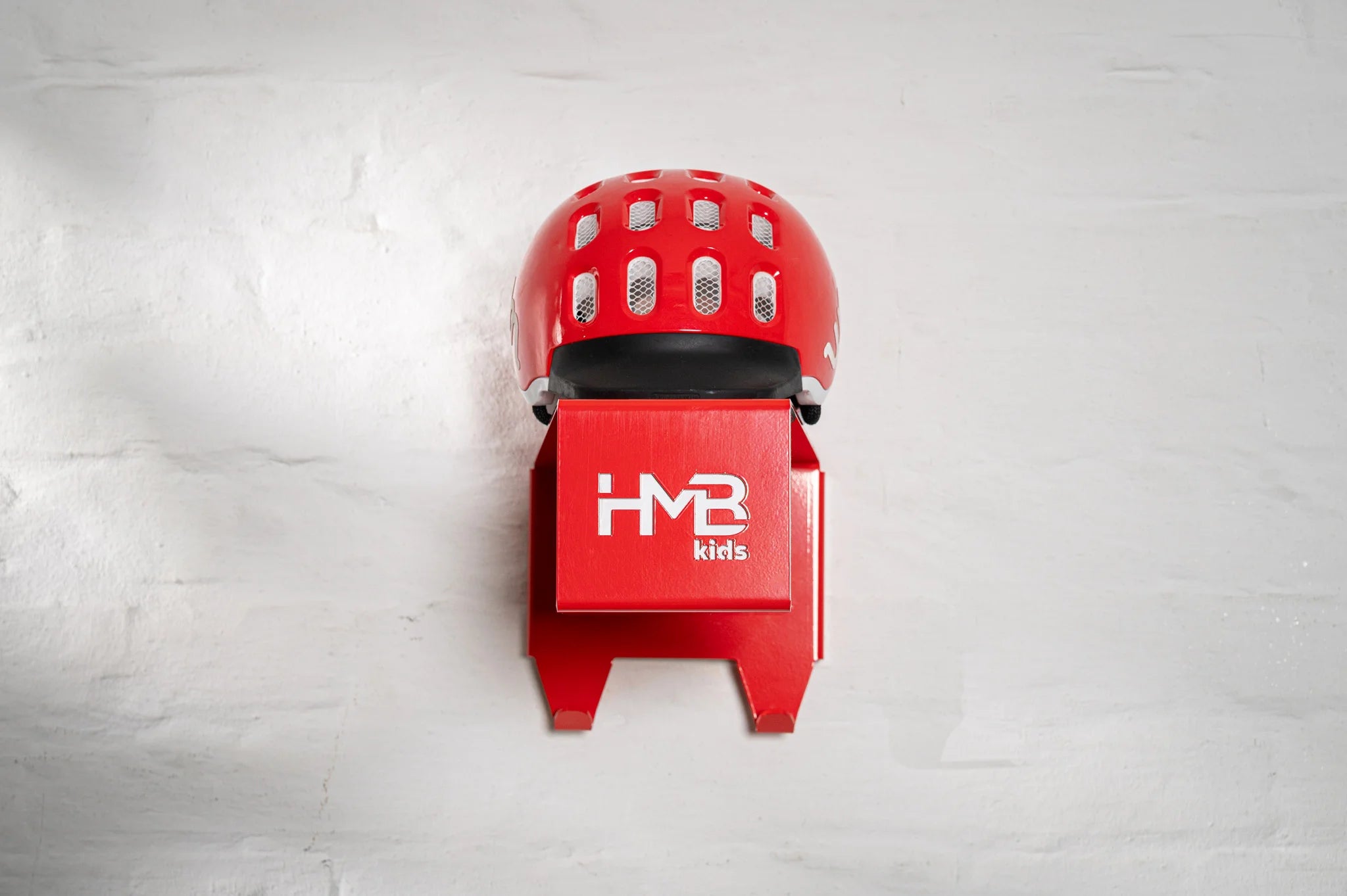 Front view of a red Hold My Bike (HMB) Kids wall mount with a matching red woom helmet resting on the top shelf, showcasing the laser-cut branding and two integrated hooks for cycling gear.