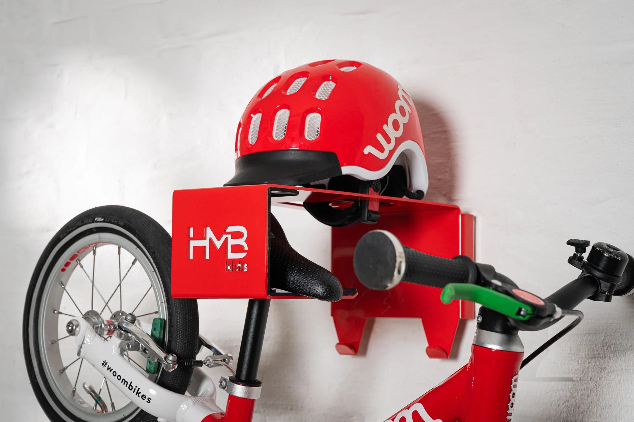 A red Hold My Bike (HMB) Kids wall mount holding a woom 1 balance bike by the saddle, with a matching red woom helmet resting on the integrated top shelf and visible lower gear hooks.