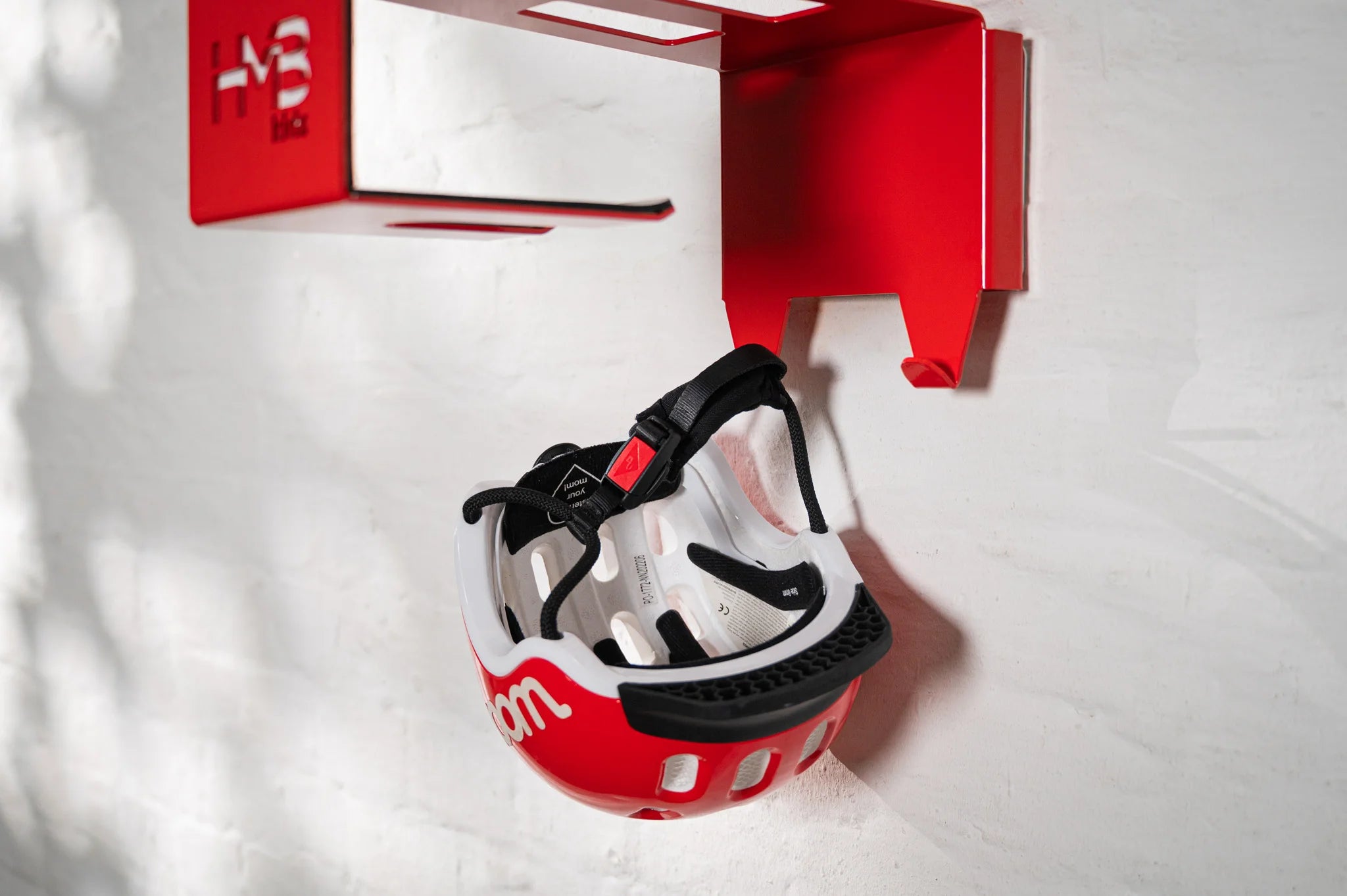 Close-up of the red Hold My Bike (HMB) Kids wall mount featuring a matching red and white woom children's helmet hanging securely by its strap
