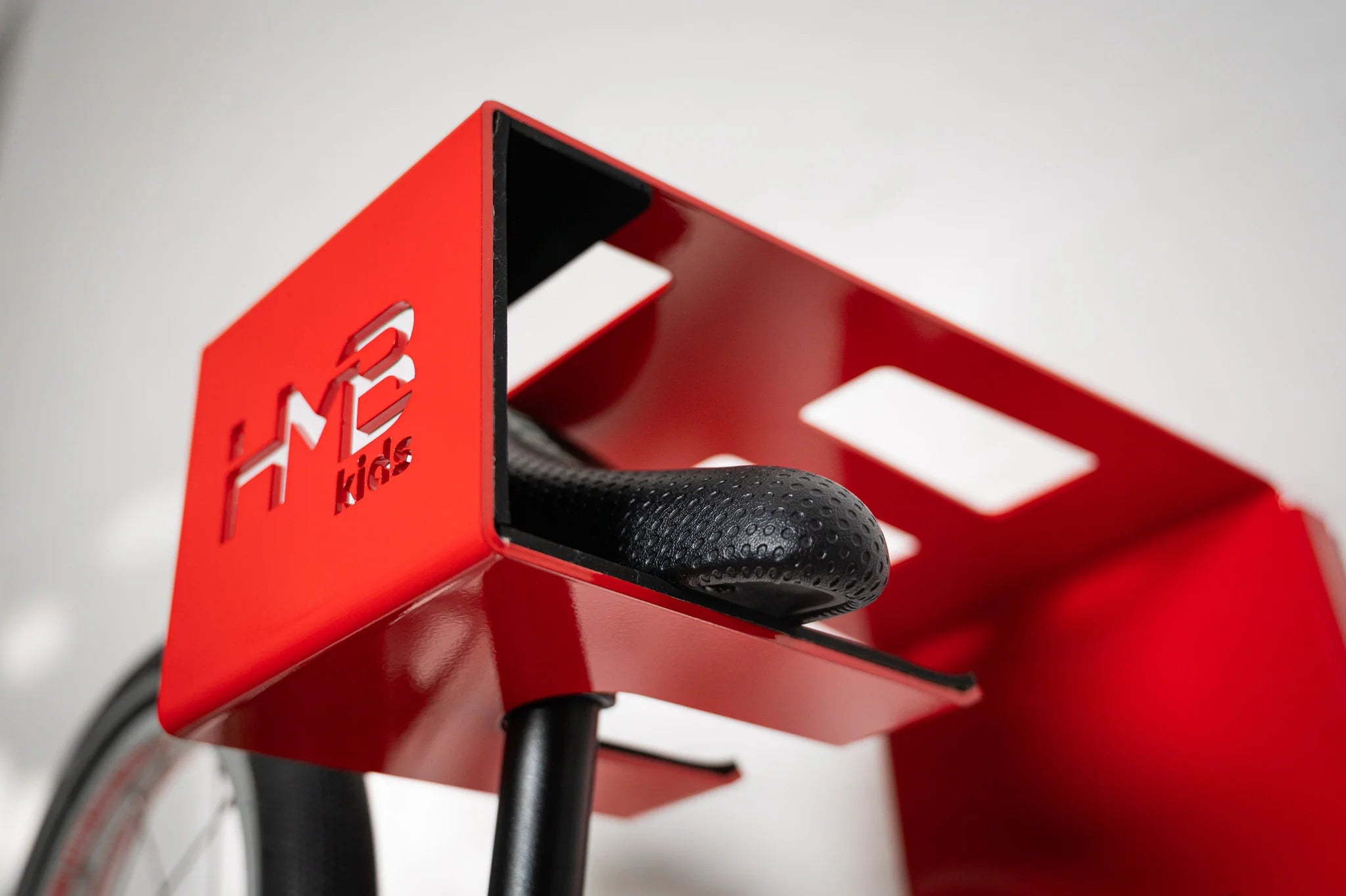 Macro view of a bicycle saddle resting securely inside the padded slot of a red Hold My Bike (HMB) Kids wall mount, featuring the laser-cut 'HMB Kids' logo and protective interior lining.