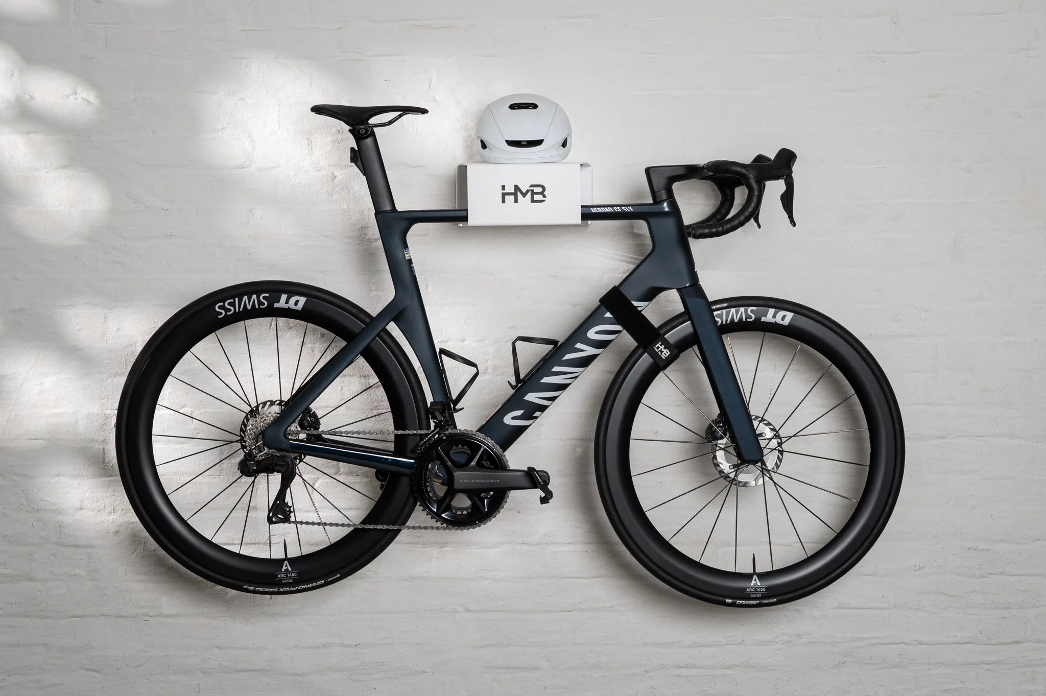A side-profile view of a matte black Canyon Aeroad CF SLX road bike perfectly displayed on a white HMB Minimal wall mount, featuring a white helmet on the top shelf and an HMB wheel strap securing the front wheel for a stable and clean presentation.