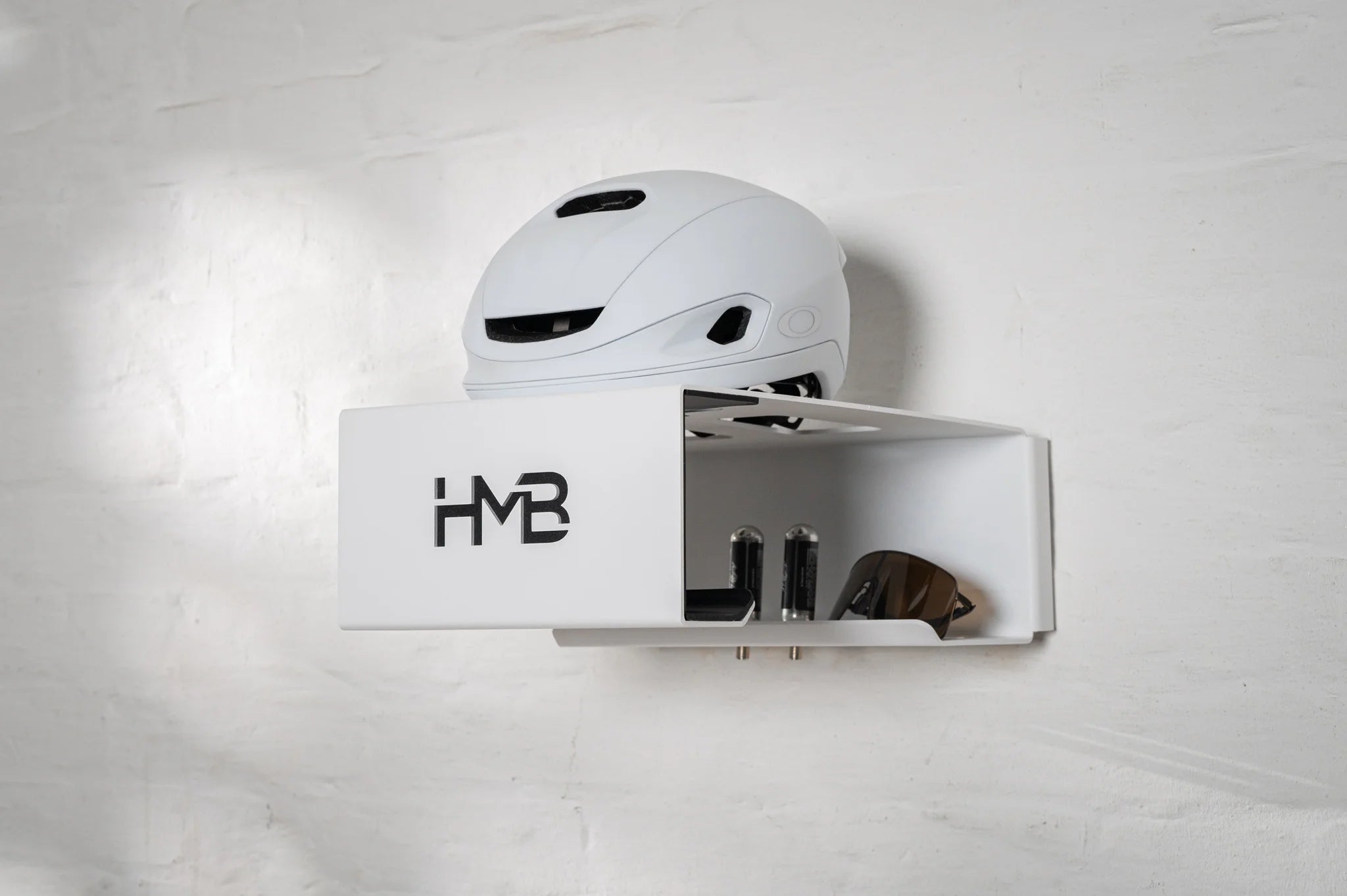 Perspective view of the white Hold my Bike (HMB) Minimal wall mount organizing a white cycling helmet, technical eyewear, and two CO2 cartridges in their dedicated storage slots against a white brick wall.
