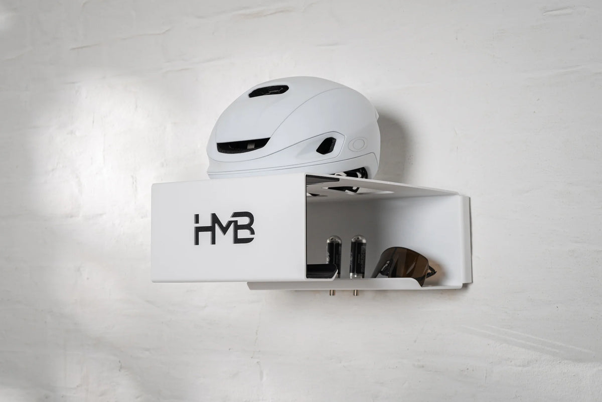 Perspective view of the white Hold my Bike (HMB) Minimal wall mount organizing a white cycling helmet, technical eyewear, and two CO2 cartridges in their dedicated storage slots against a white brick wall.