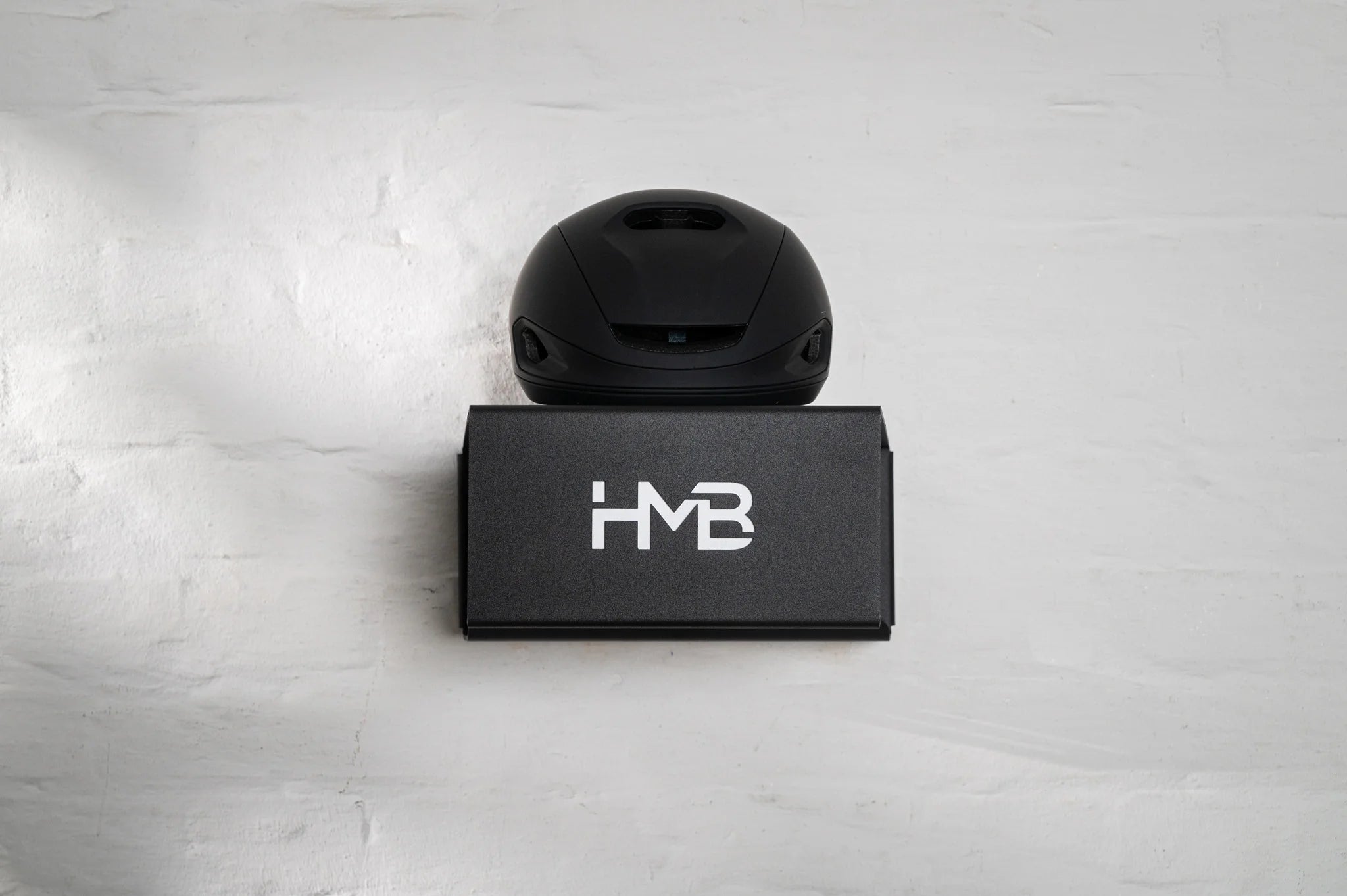 A front-facing, minimalist view of the black HMB wall mount installed on a white brick wall, featuring a matte black cycling helmet on the top shelf and the signature laser-cut 'HMB' logo on the front panel.