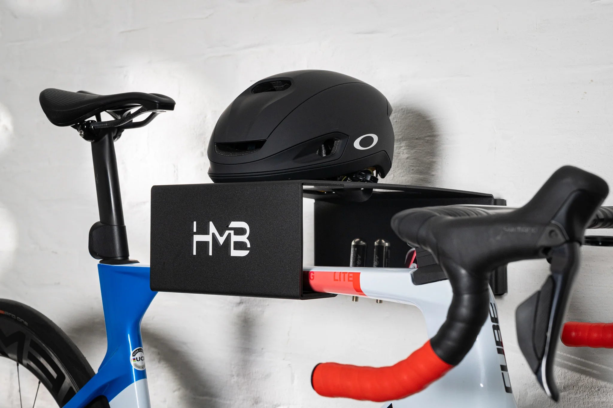 Close-up of a blue and white Cube road bike stored on the black HMB Ultimate wall mount, showing a black helmet on the top shelf and integrated storage slots holding two CO2 cartridges for ride-ready organization.