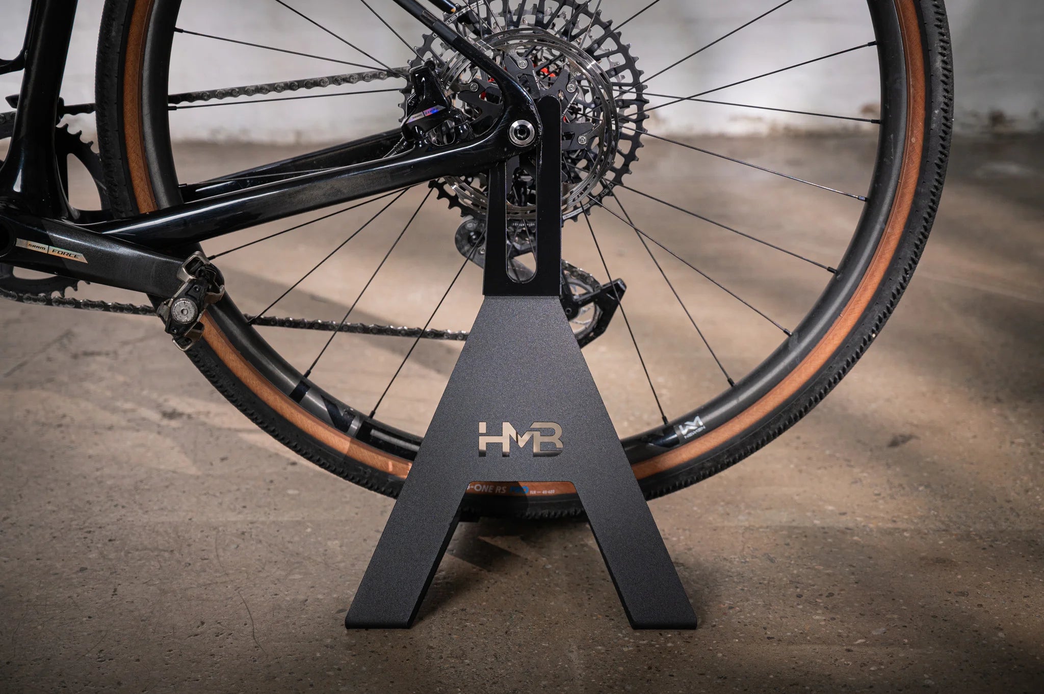 Close-up of the black Hold My Bike (HMB) bike stand, securely holding the rear wheel of a gravel bike by the thru-axle without contacting the frame or rear derailleur.