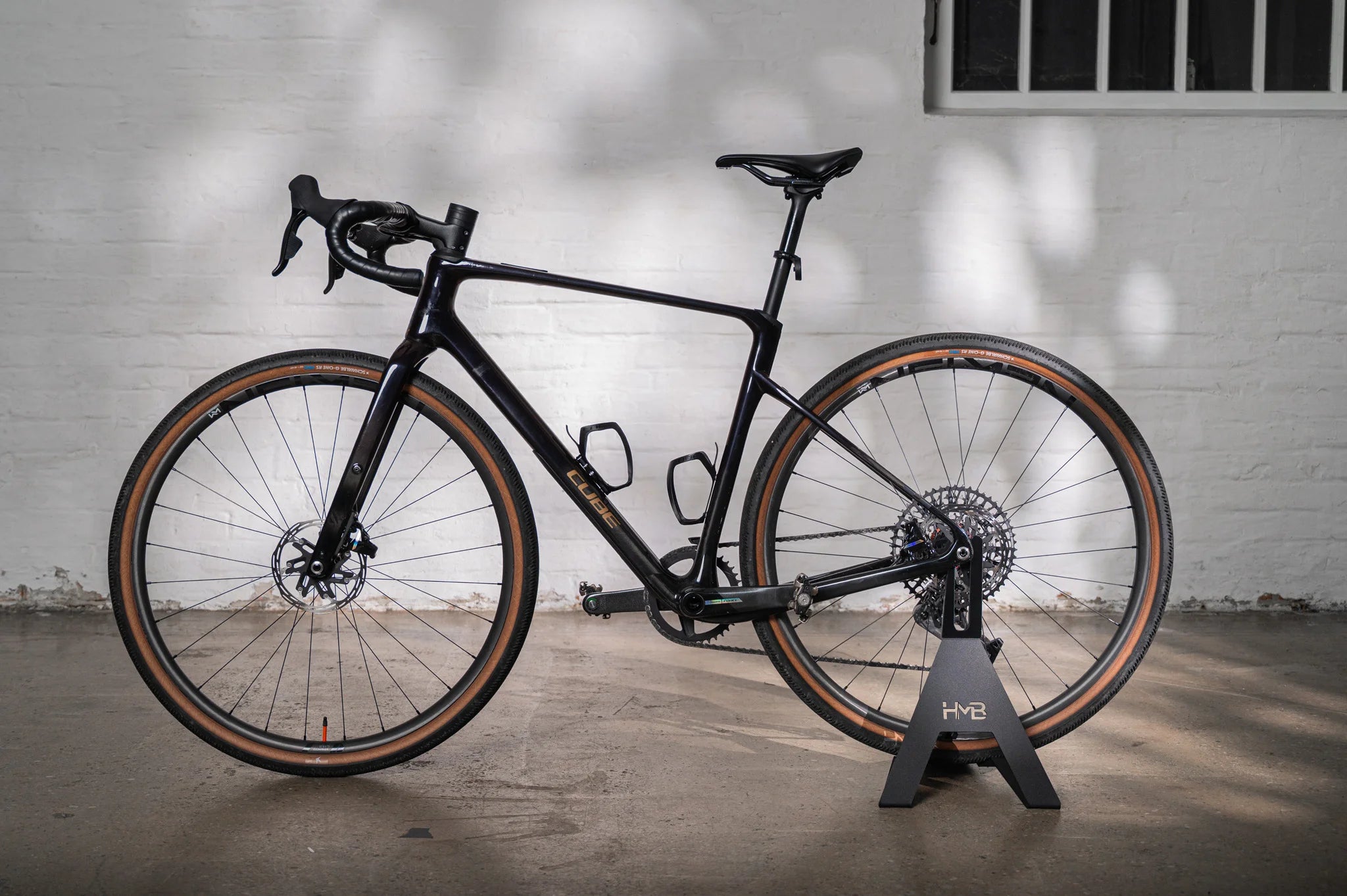 A black Cube gravel bike with classic tan-wall tires, displayed in the sleek black Hold My Bike (HMB) bike stand against a light brick wall.