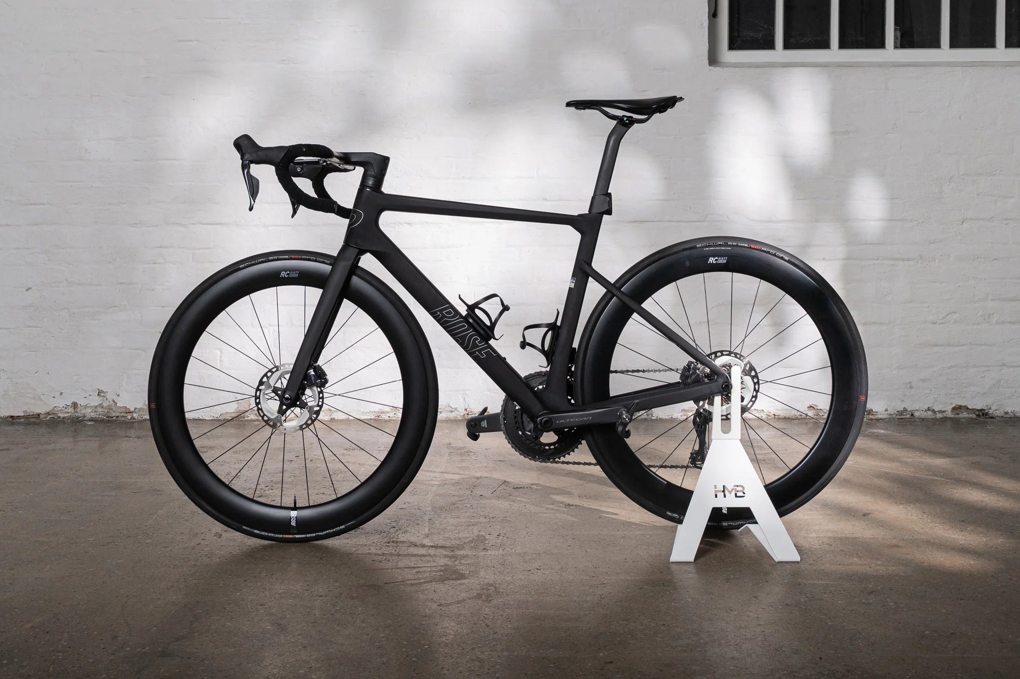 A matte black Rose XLITE 06 road bike displayed in the minimalist white Hold My Bike (HMB) bike stand against a light brick wall.