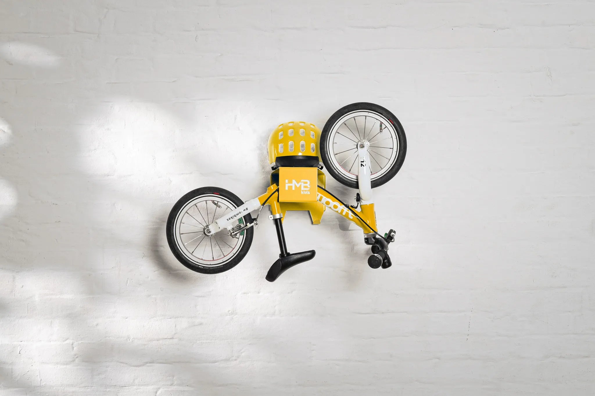 A yellow Hold My Bike (HMB) Kids wall mount displaying a yellow woom 1 balance bike in a vertical storage position, with a matching yellow woom helmet resting securely on the top shelf.