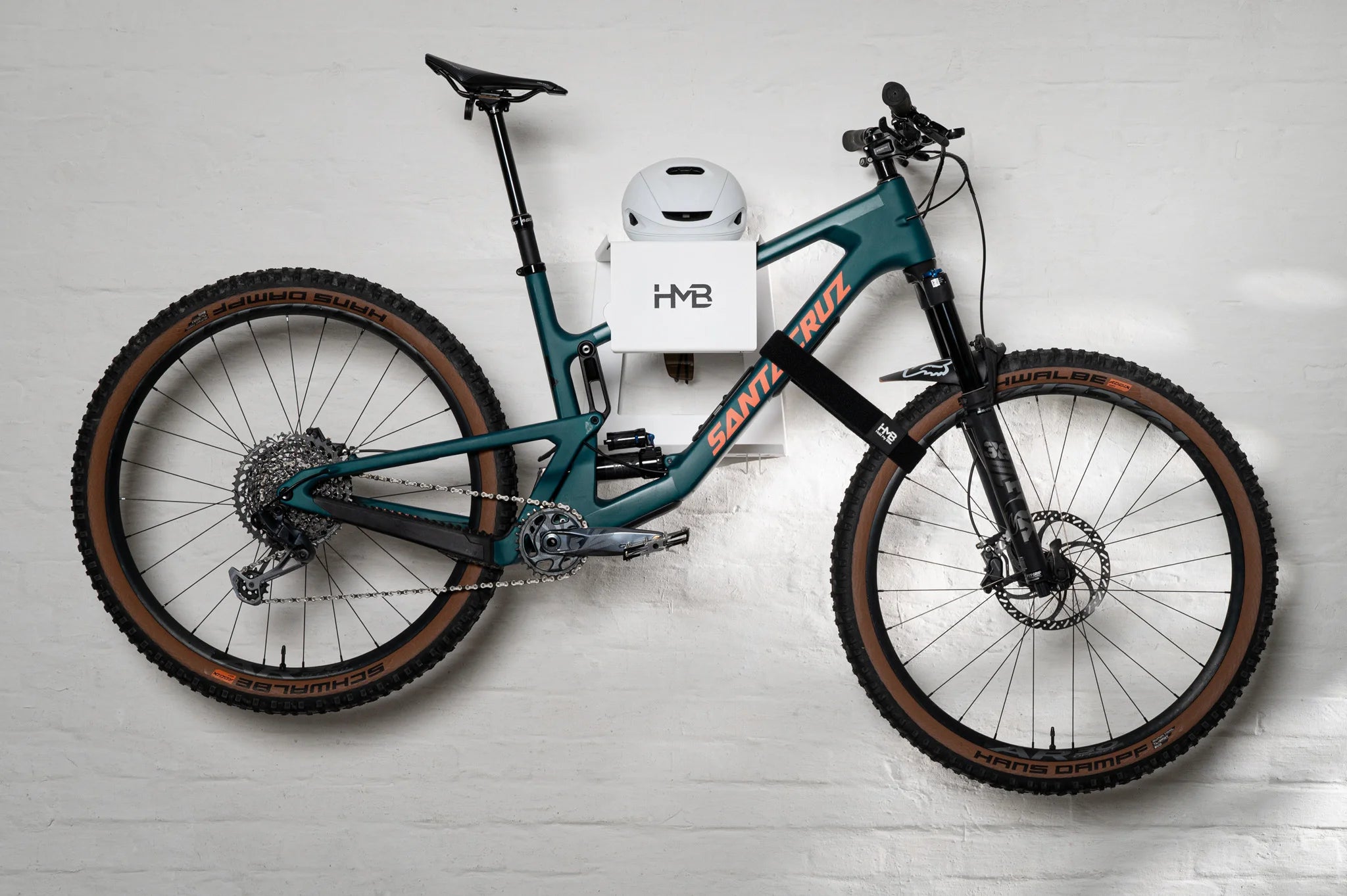 Wide shot of a teal Santa Cruz mountain bike perfectly leveled on a white HMB Ultimate XL wall mount, featuring an integrated white helmet on the top shelf and a black HMB wheel strap securing the front wheel against a white brick wall.