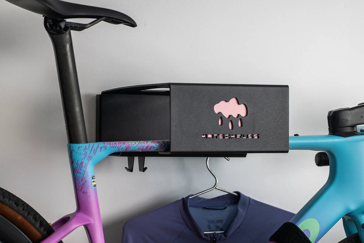 A black Hold My Bike (HMB) CUSTOM wall mount with a pink customised Matschfuss by Plattfuss Podcast front plate displaying a light blue and pink Tavelo Grow gravel bike with a purple Ryzon Signature Series jersey on a silver metal hanger.