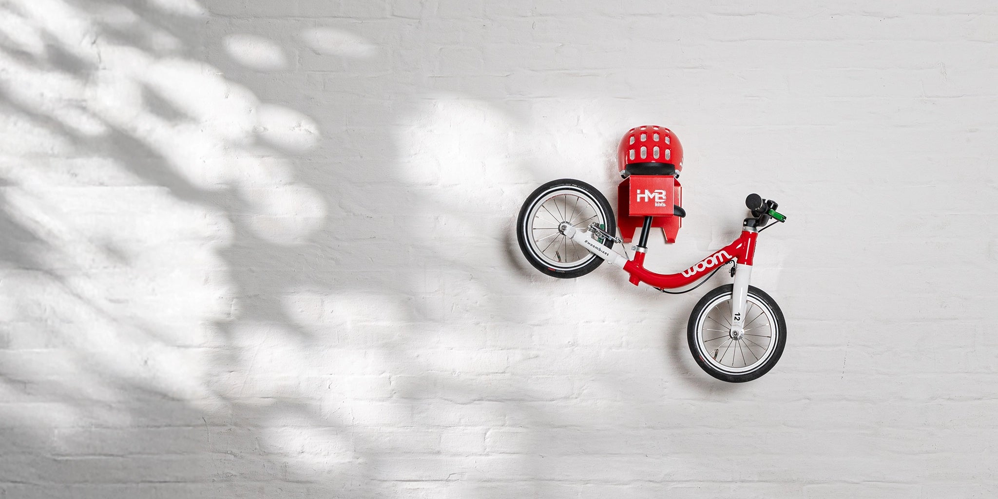 A wide lifestyle shot of a red woom 1 balance bike and matching helmet neatly organized on a red Hold My Bike (HMB) Kids wall mount, centered on a white brick wall with natural light and soft leaf shadows.