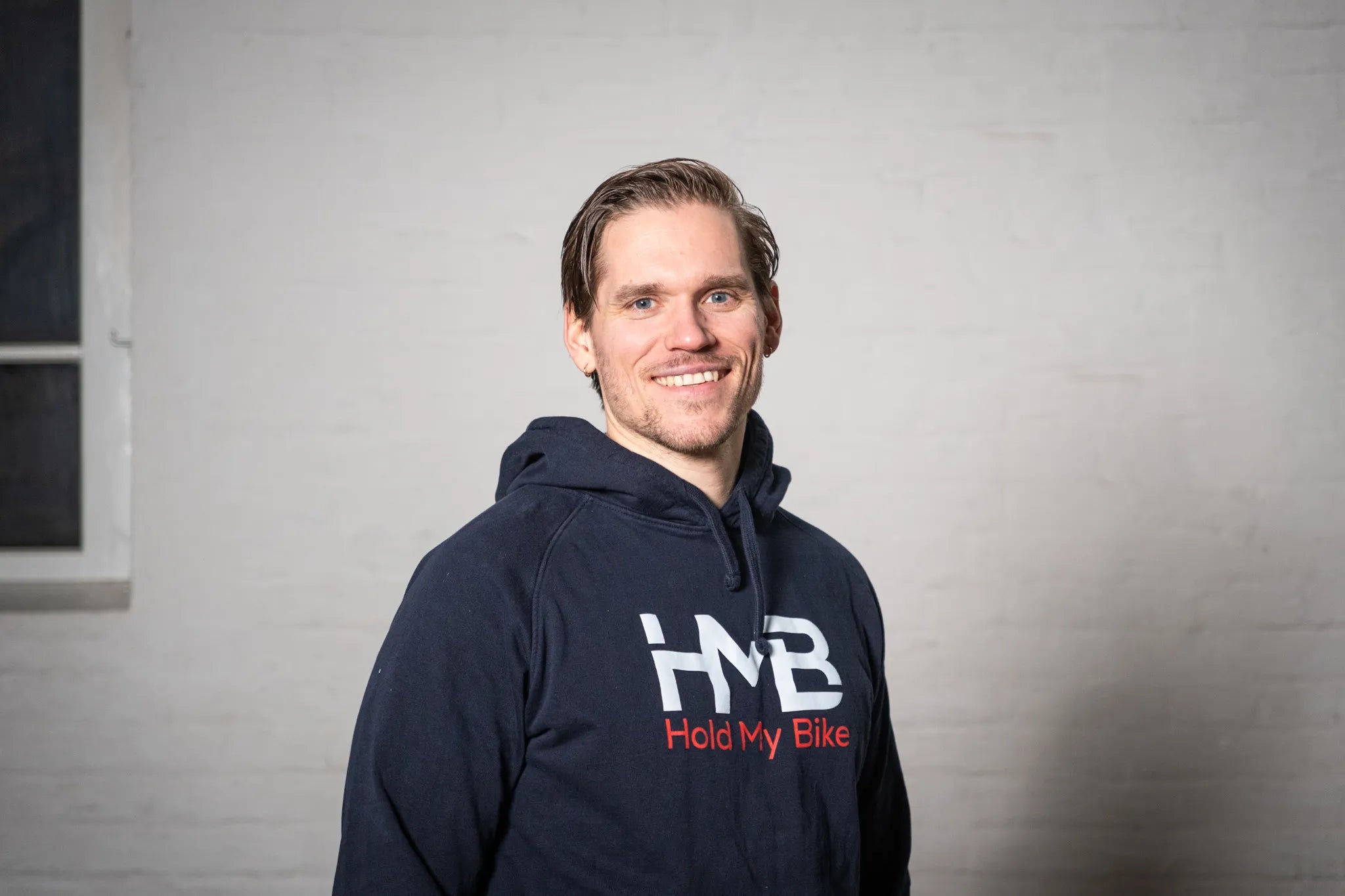 Hold My Bike (HMB) founder Kai Fields wearing branded hoodies in front of a white brick wall – experts in bike wall mounts.