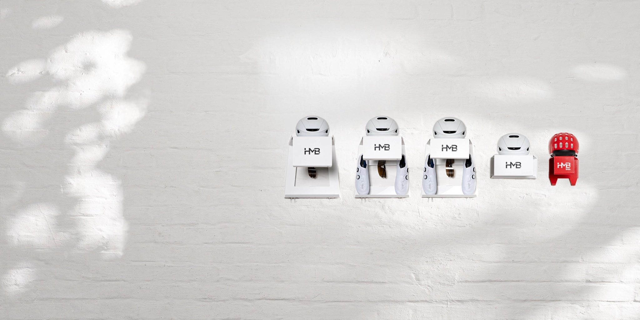 Minimalist desktop banner of the Hold My Bike (HMB) portfolio against a white brick wall: Displaying various mounting systems in white and red for bikes, helmets, and shoes, combining organization with modern home design.