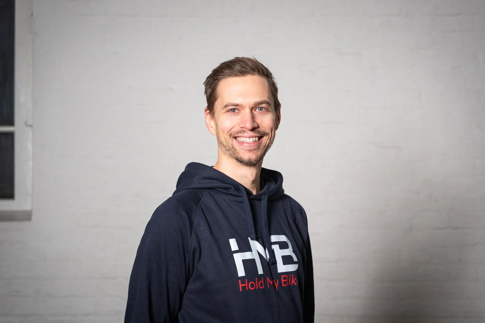 Hold My Bike (HMB) founder Mathias Guggenbichler wearing branded hoodies in front of a white brick wall – experts in bike wall mounts.