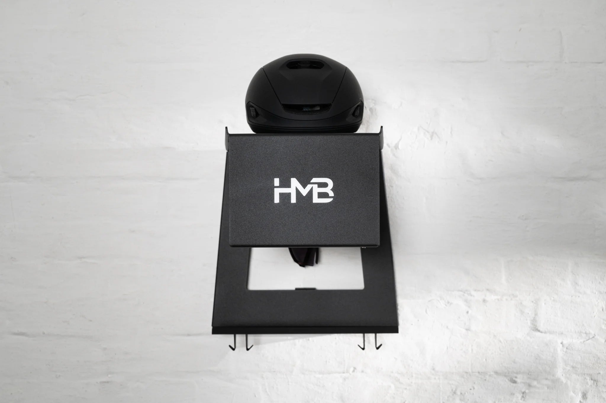 Front-facing view of the matte black HMB Ultimate wall mount with a black cycling helmet on the top shelf, showcasing the laser-cut 'HMB' logo and integrated lower hooks for gear organization.