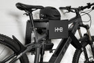 Side-profile view of a Trek E-MTB secured to the black HMB Ultimate XL wall mount, highlighting the heavy-duty frame cradle, integrated helmet shelf, and the use of an HMB wheel strap to keep the front wheel perfectly straight.
