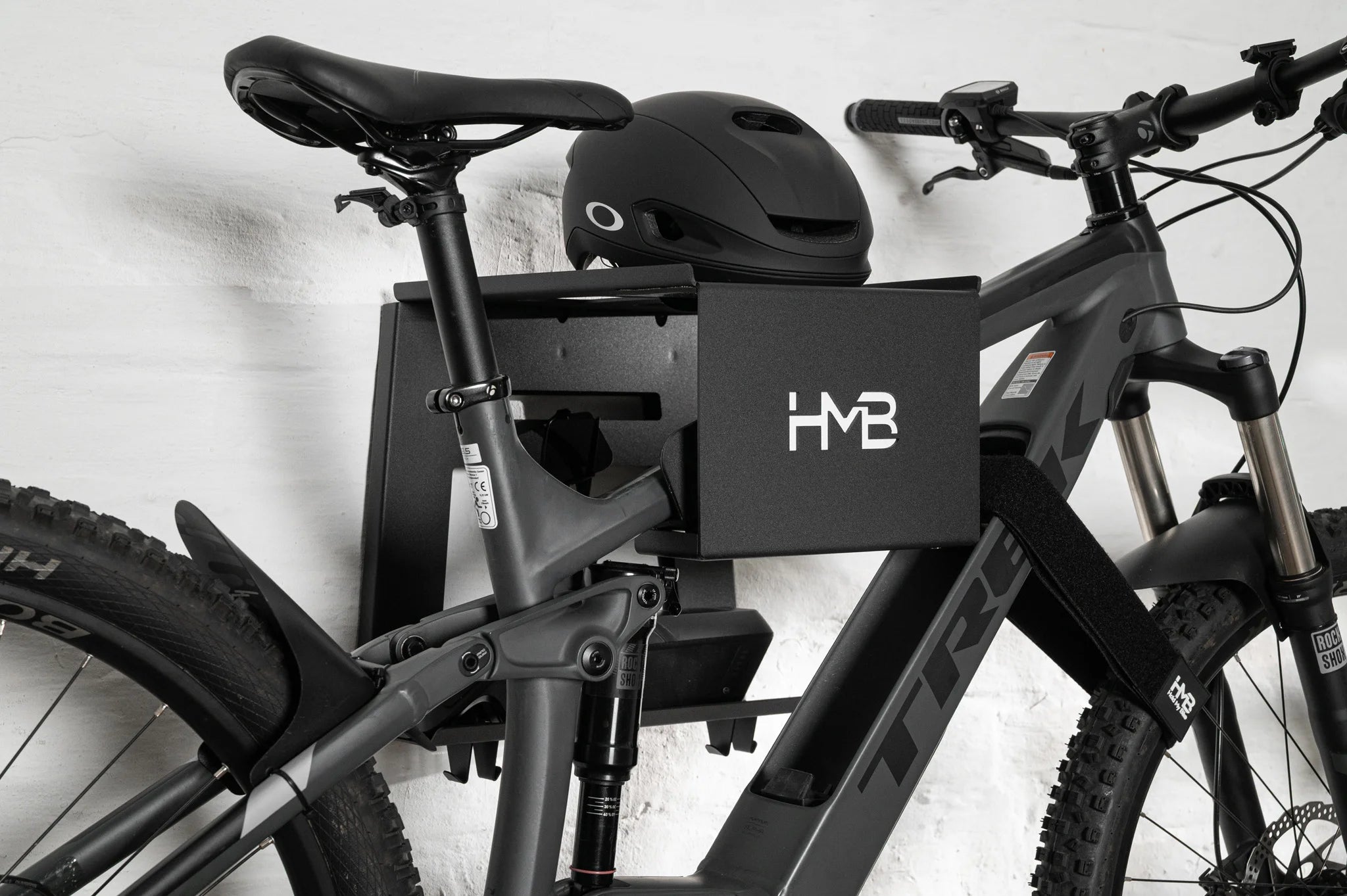 Side-profile view of a Trek E-MTB secured to the black HMB Ultimate XL wall mount, highlighting the heavy-duty frame cradle, integrated helmet shelf, and the use of an HMB wheel strap to keep the front wheel perfectly straight.