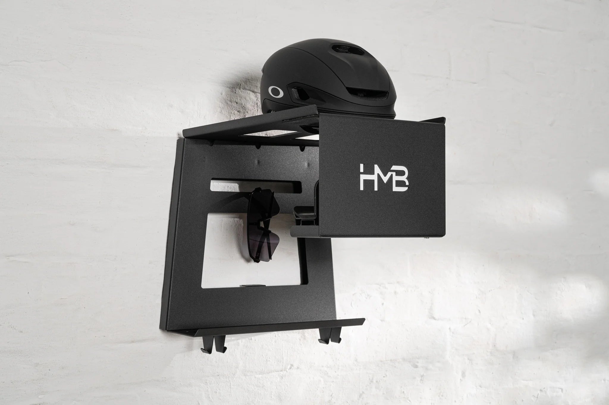 Perspective view of the black HMB Ultimate wall mount organizing a black Oakley cycling helmet on the top shelf and performance sunglasses in the integrated dock, mounted on a white brick wall.