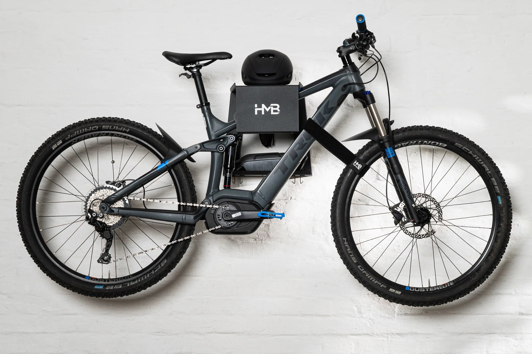 Wide shot of a Trek E-MTB secured to the black HMB Ultimate XL wall mount, showing the combined use of the integrated helmet shelf, gear storage, and an HMB wheel strap to stabilize the front wheel.