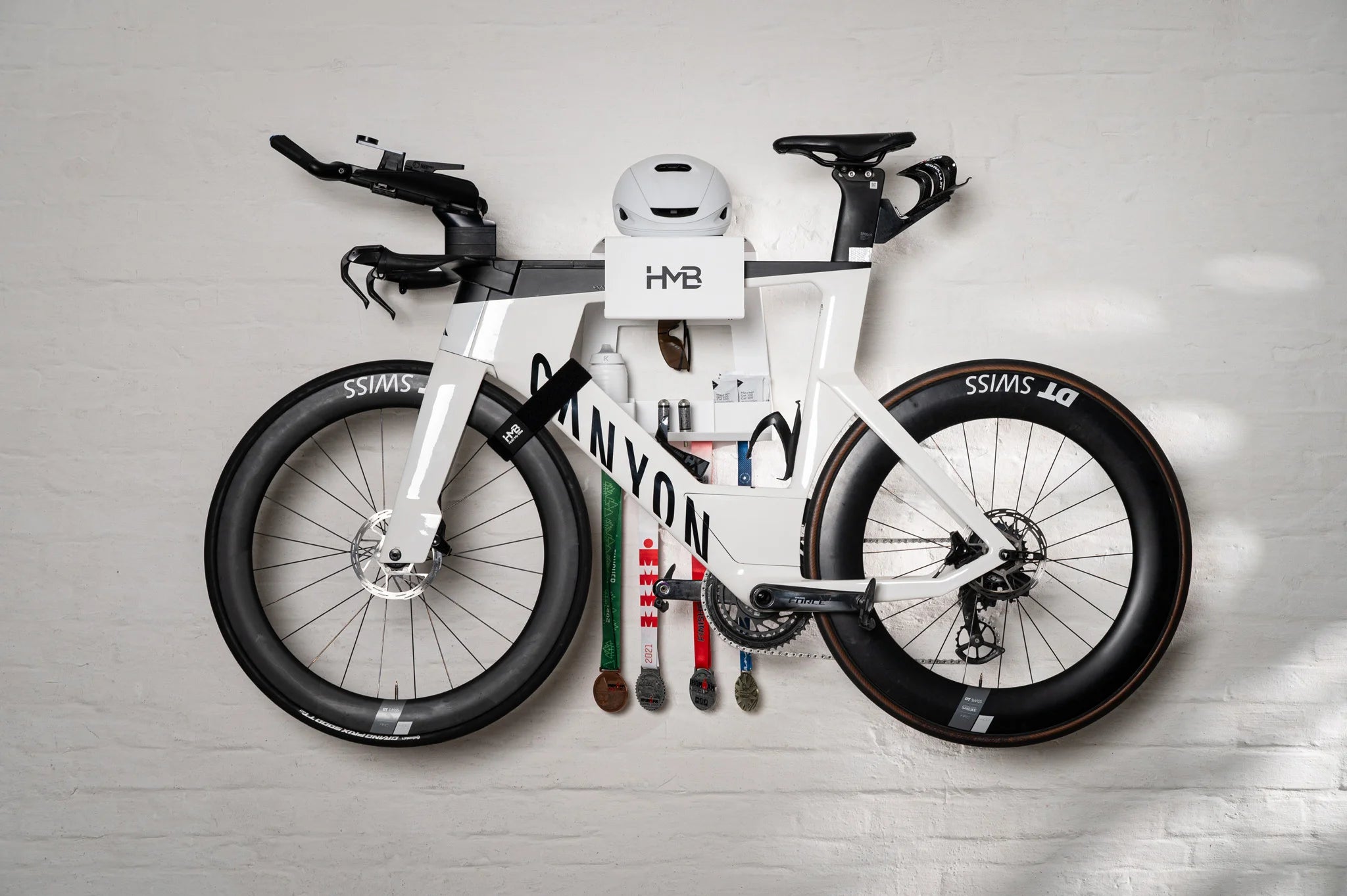 The Hold My Bike (HMB) Ultimate set in white: A Canyon Speedmax triathlon bike perfectly leveled with the innovative HMB tilt mechanism, featuring organized storage for a helmet, race medals, and CO2 cartridges.