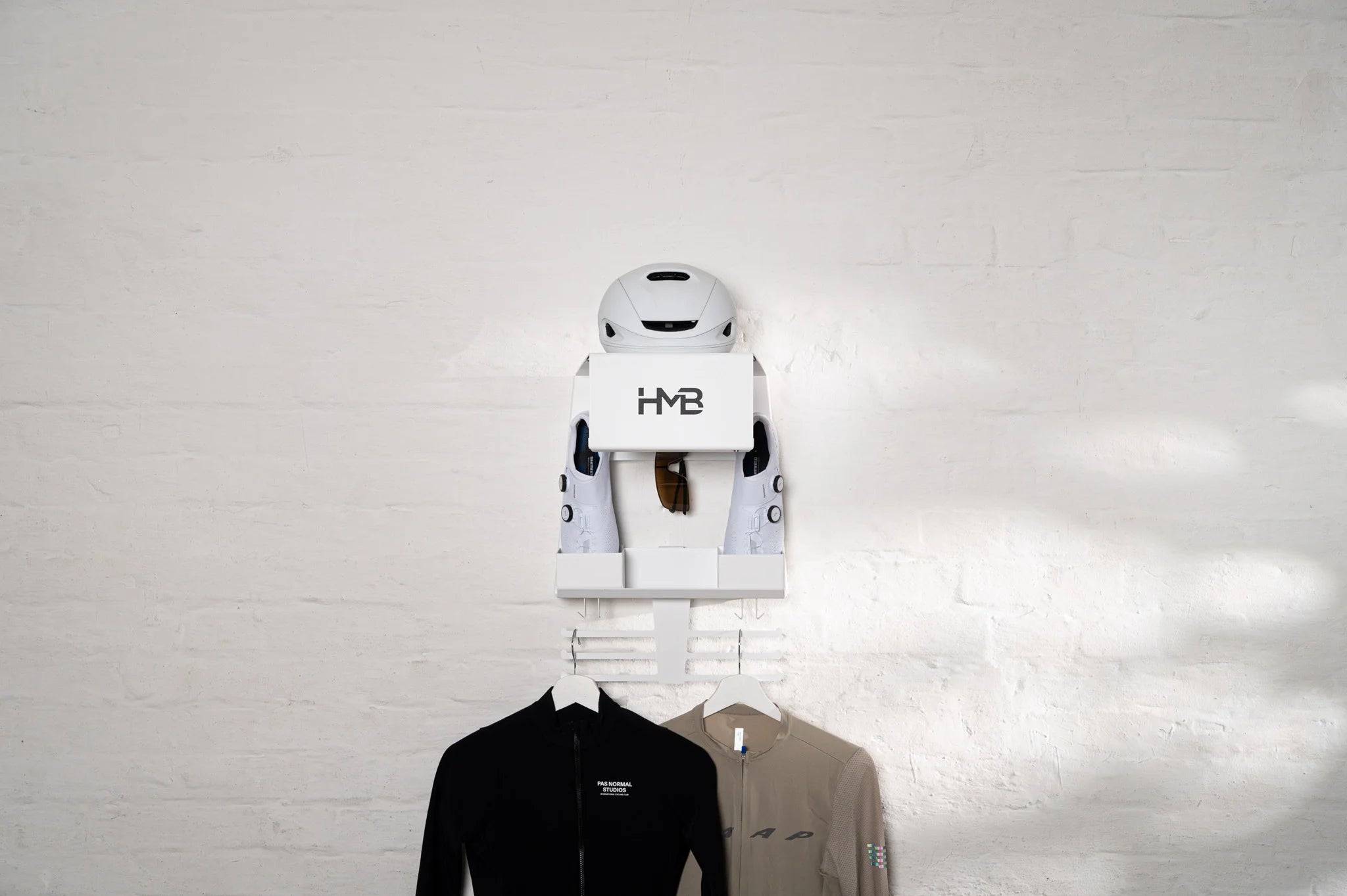 A minimalist lifestyle shot of the white Hold My Bike (HMB) Ultimate wall organizer on a white brick wall, featuring an integrated helmet shelf, a dedicated eyewear dock, vertical slots for cycling shoes, and a lower rack with jerseys on hangers.