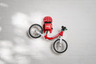 A red Hold My Bike (HMB) Kids wall mount holding a red woom 1 balance bike by the seat post, with a matching red woom helmet resting on the integrated top shelf.