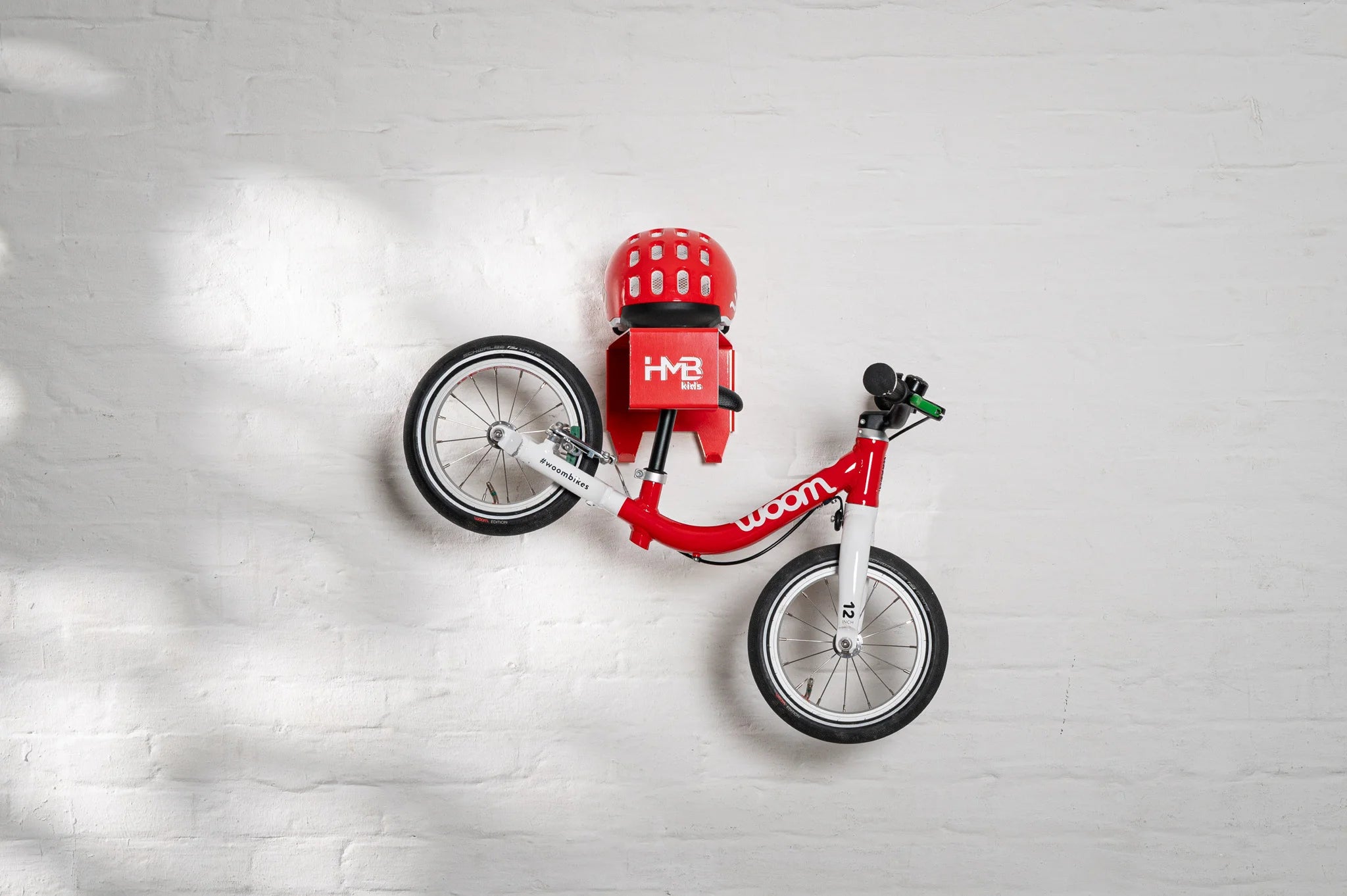 A red Hold My Bike (HMB) Kids wall mount holding a red woom 1 balance bike by the seat post, with a matching red woom helmet resting on the integrated top shelf.
