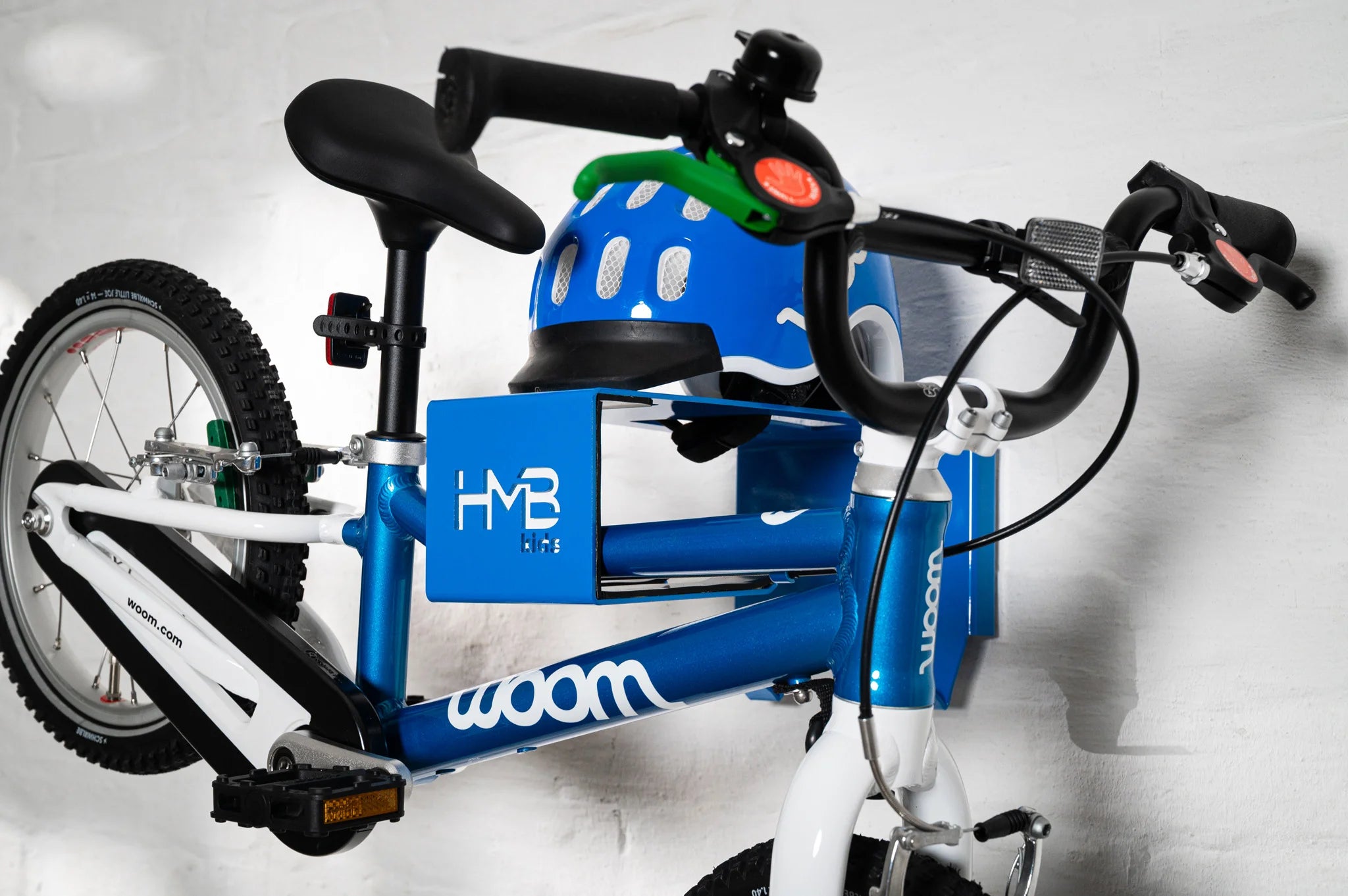 A blue Hold My Bike (HMB) Kids wall mount holding a blue woom 2 children's bike by the frame, featuring an integrated top shelf with a matching blue woom helmet and visible lower hooks for gear storage.