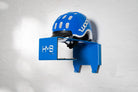 Perspective view of the blue HMB Kids wall mount with a blue helmet, showcasing the laser-cut branding and dual lower hooks for hanging cycling accessories.