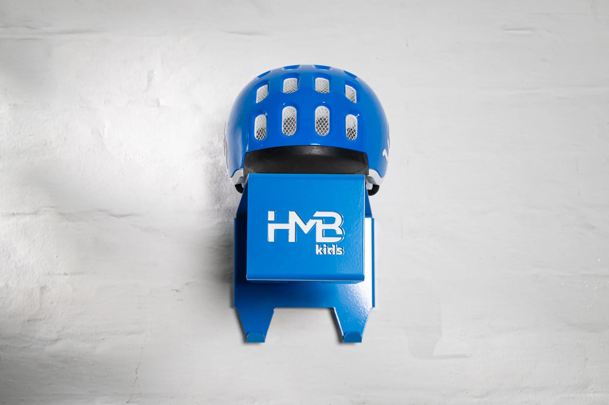 Front view of a blue Hold My Bike (HMB) Kids wall mount with a matching blue woom helmet resting on the integrated top shelf, showcasing the laser-cut 'HMB kids' logo and dual lower hooks.