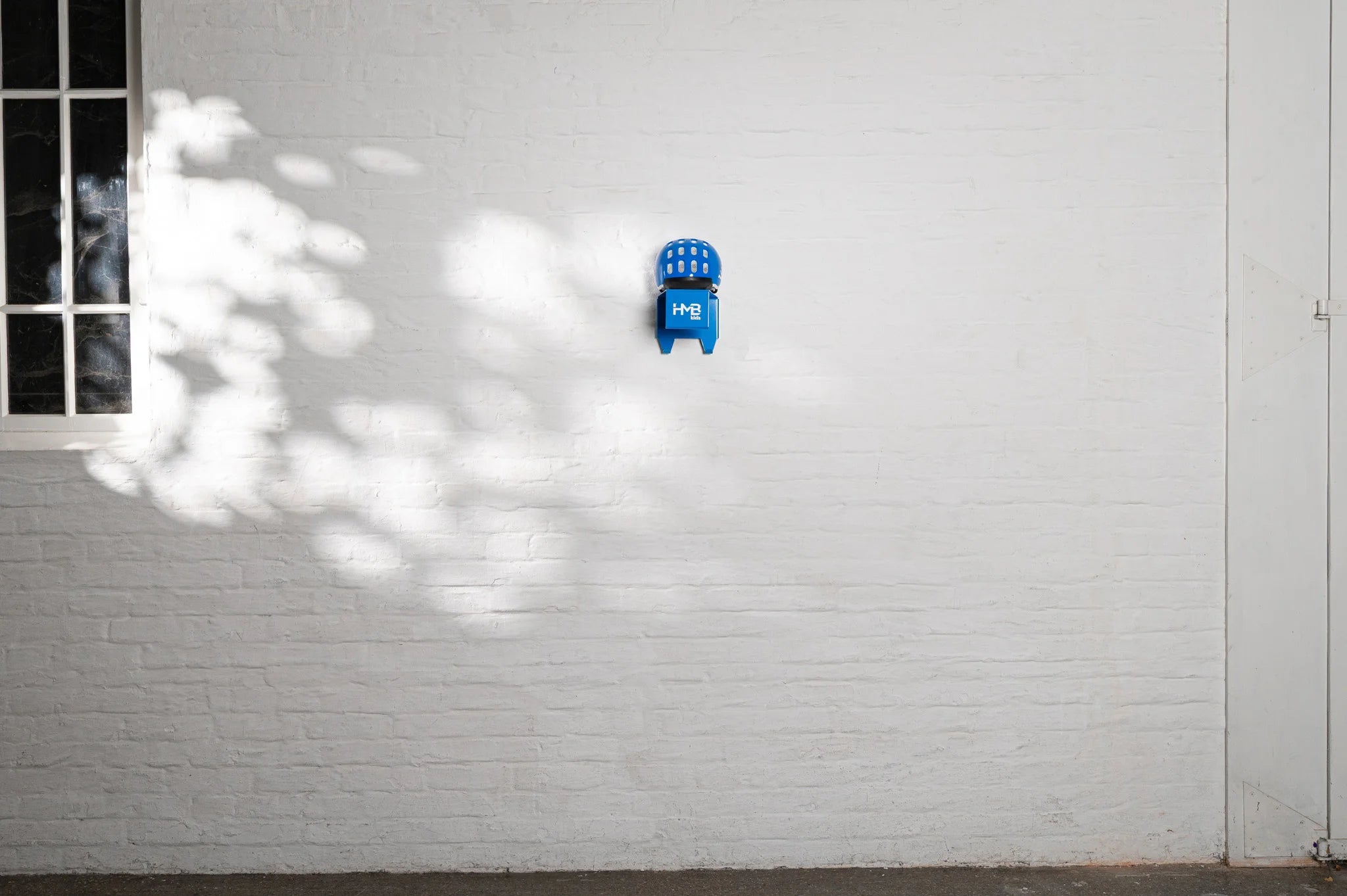 A blue woom helmet organized on a blue Hold My Bike (HMB) Kids wall mount, showcasing a clean and modern storage solution for children's cycling gear against a white brick wall.