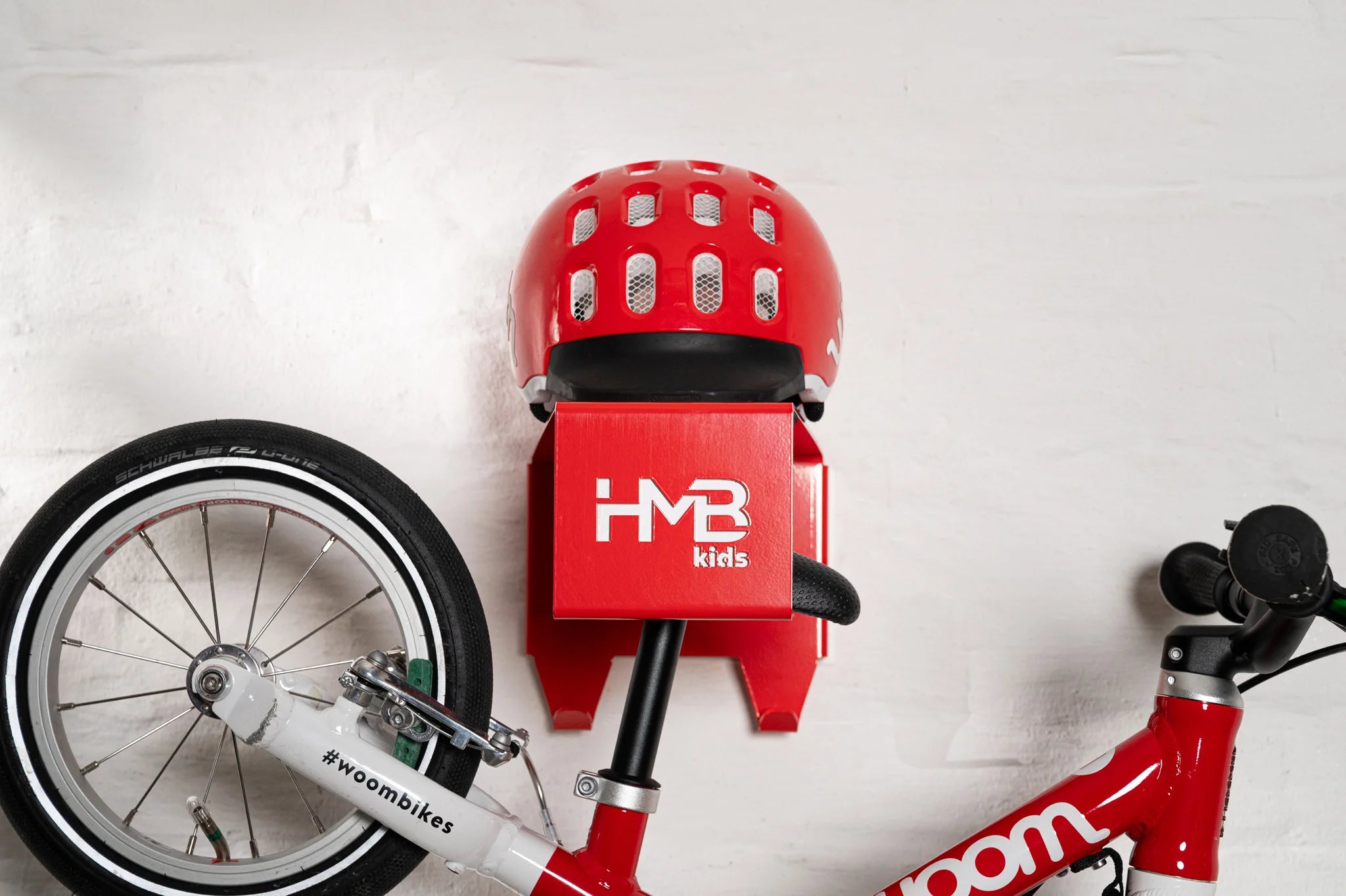 Close-up of a red Hold My Bike (HMB) Kids wall mount holding a red woom 1 balance bike by the seat post, with a matching red woom helmet resting on the integrated top shelf against a white brick wall.