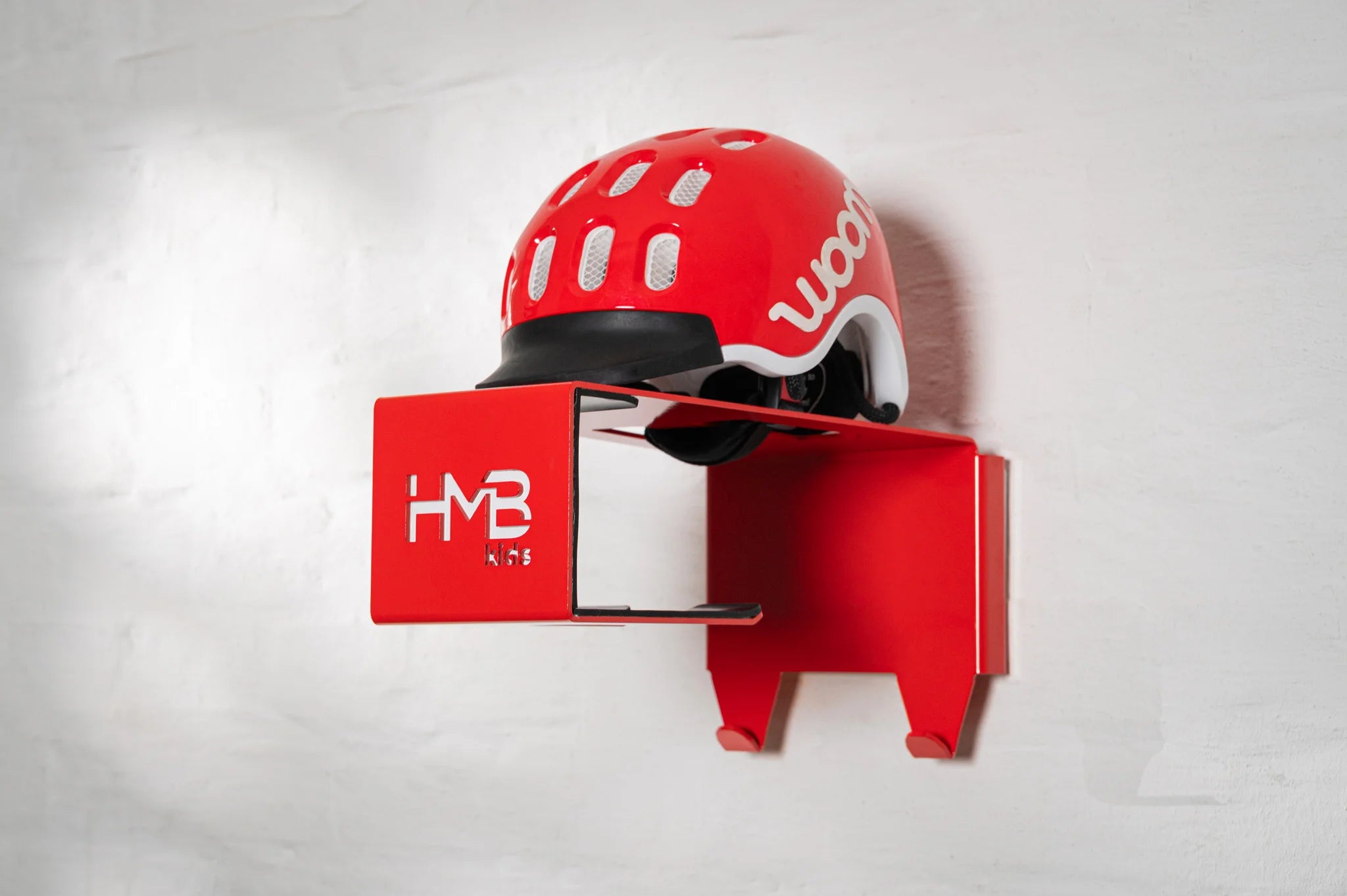Perspective view of a red Hold My Bike (HMB) Kids wall mount with a matching red woom helmet on the top shelf, highlighting the laser-cut 'HMB Kids' branding and dual integrated equipment hooks.