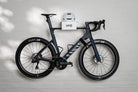 A side-profile view of a matte black Canyon Aeroad CF SLX road bike perfectly displayed on a white HMB Minimal wall mount, featuring a white helmet on the top shelf and an HMB wheel strap securing the front wheel for a stable and clean presentation.
