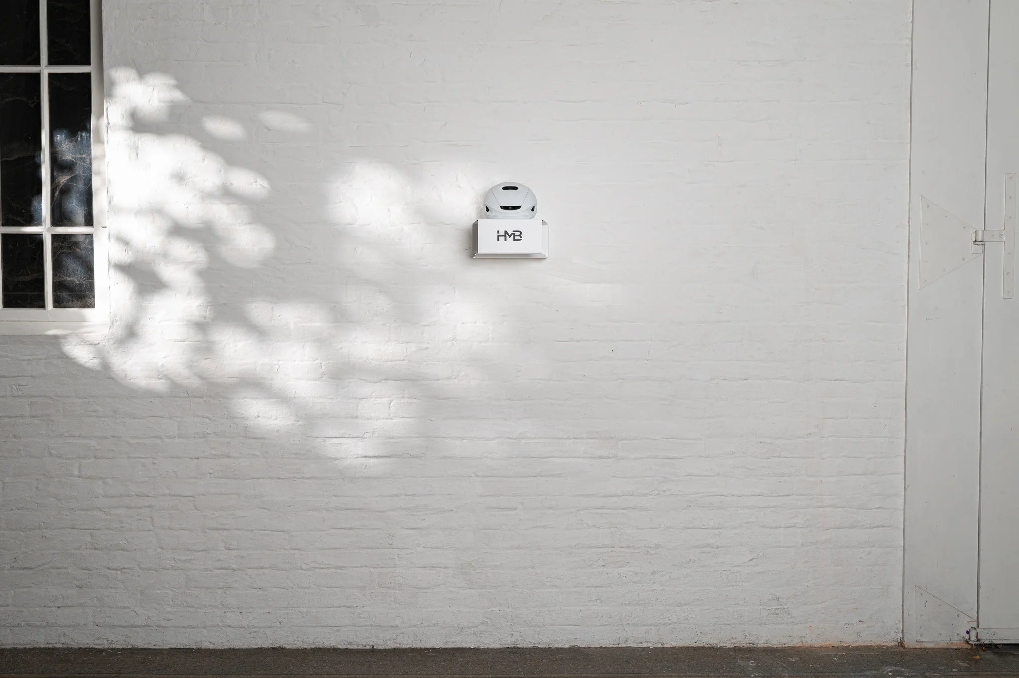 Wide architectural shot of a white HMB Minimal wall mount installed on a white brick wall in a naturally lit room, featuring a white cycling helmet on the top shelf and highlighting its sleek, low-profile design.