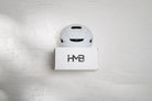 A front-facing view of the white HMB Minimal wall mount installed on a white brick wall, featuring a white cycling helmet on the top shelf and the signature laser-cut 'HMB' logo on the front panel.