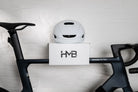A front-facing view of a matte black Canyon Aeroad CF SLX road bike securely stored on a white HMB Minimal wall mount, featuring a white cycling helmet on the top shelf against a white brick wall.