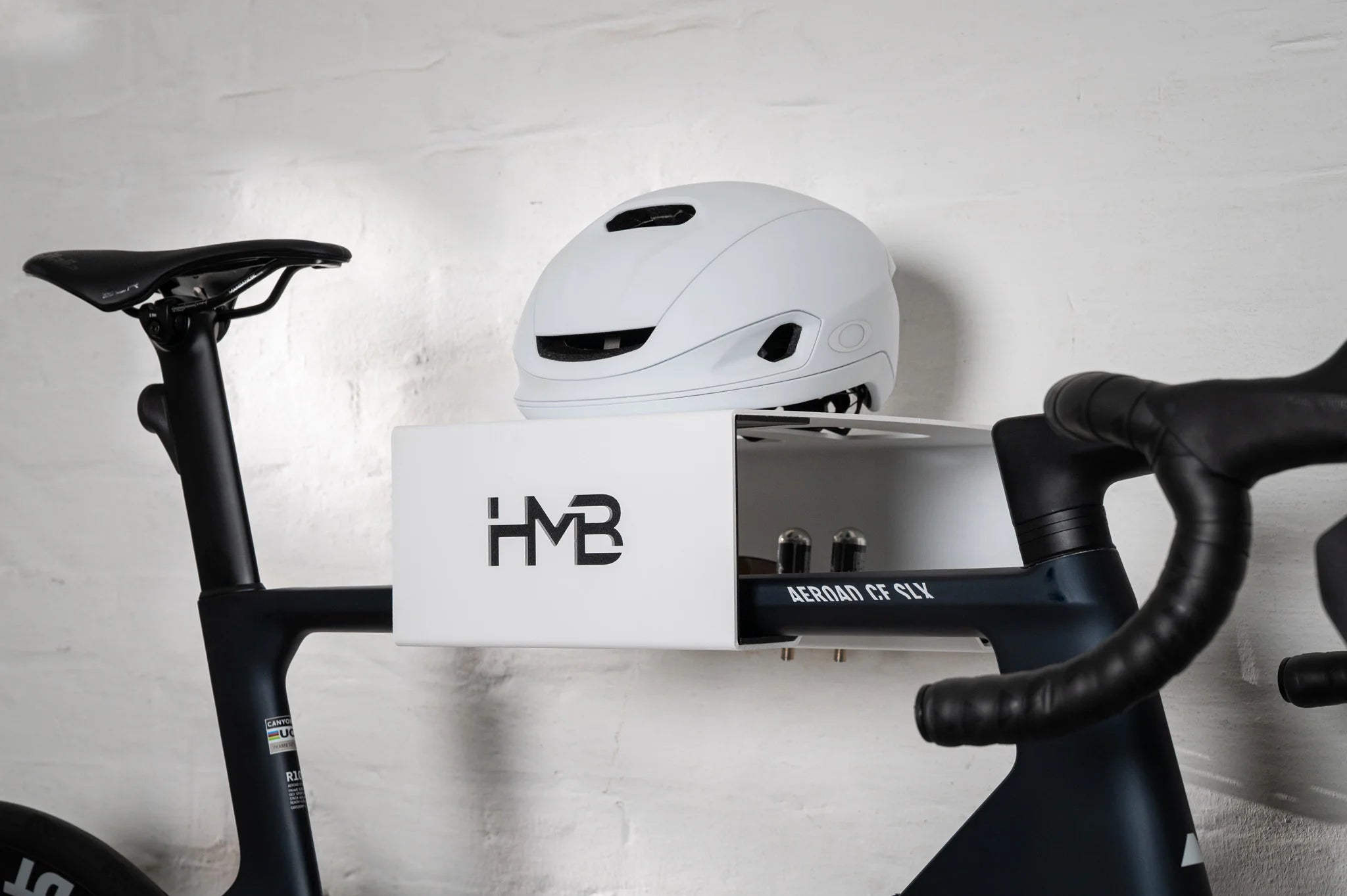A close-up view of a matte black Canyon Aeroad CF SLX road bike stored on a white HMB Minimal wall mount, featuring a white helmet on the top shelf and integrated slots holding two CO2 cartridges.