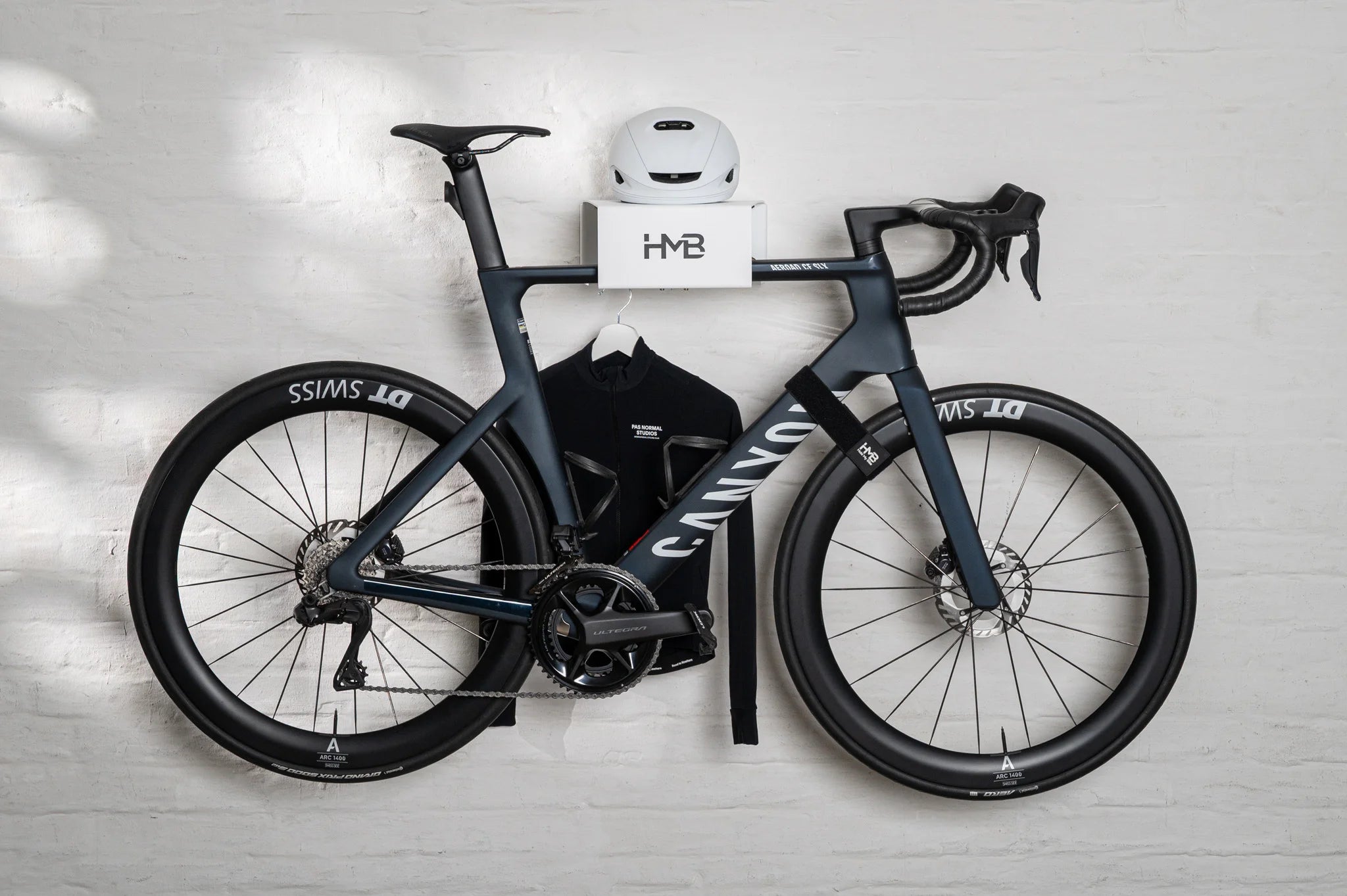 A black Canyon road bike stored on the white HMB minimal wall mount, featuring a top shelf for a helmet and an integrated lower hanger for cycling apparel, secured against a white brick wall.
