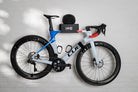 A side-profile shot of a blue and white Cube aero road bike mounted on the black HMB Ultimate wall mount, featuring an integrated helmet on the top shelf and an HMB wheel strap securing the front wheel to the frame for a clean, stable display.