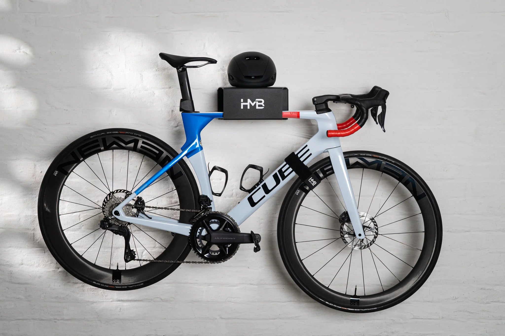 A side-profile shot of a blue and white Cube aero road bike mounted on the black HMB Ultimate wall mount, featuring an integrated helmet on the top shelf and an HMB wheel strap securing the front wheel to the frame for a clean, stable display.