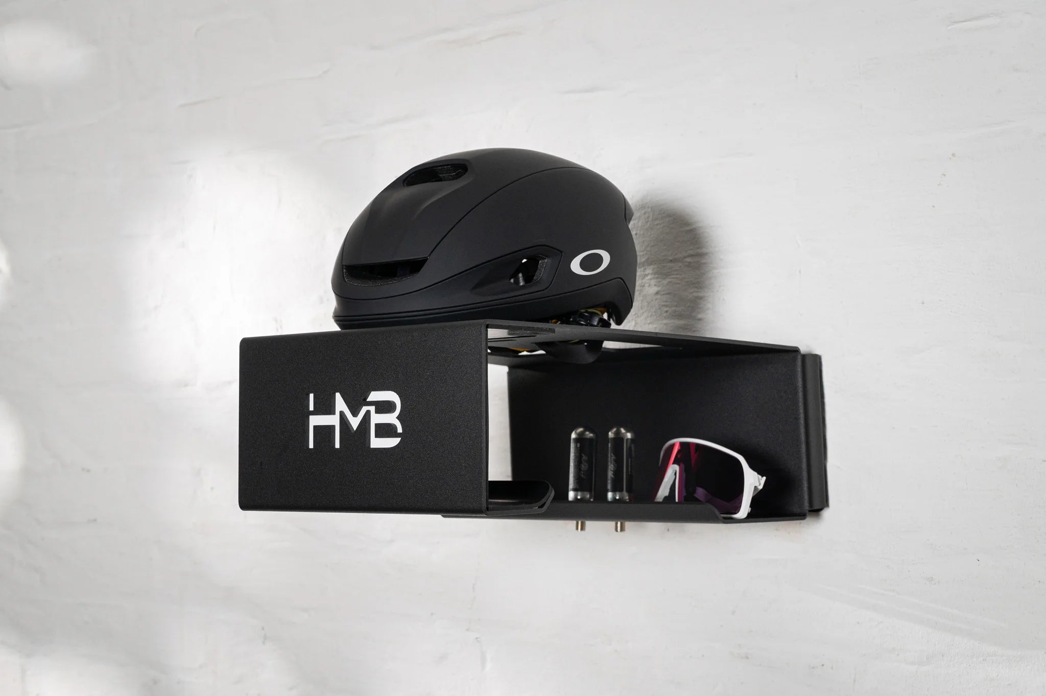 Perspective view of the black HMB Ultimate wall mount organizing a black Oakley helmet, cycling sunglasses, and two CO2 cartridges in their dedicated storage slots, mounted on a white brick wall.