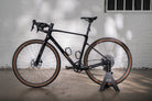 A black Cube gravel bike with classic tan-wall tires, displayed in the sleek black Hold My Bike (HMB) bike stand against a light brick wall.